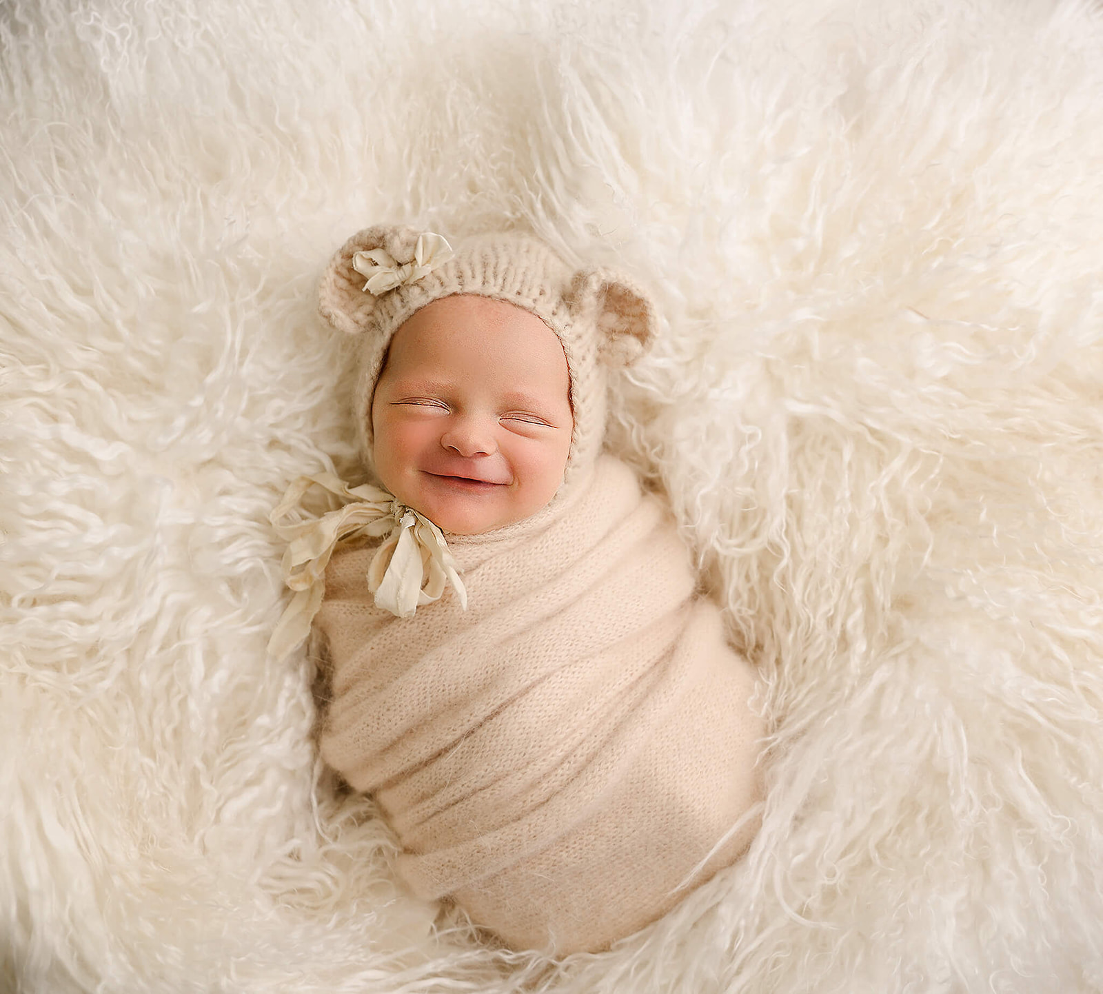 A smiling newborn baby sleeps in a swaddle and knit hat with ears on a furry blanket