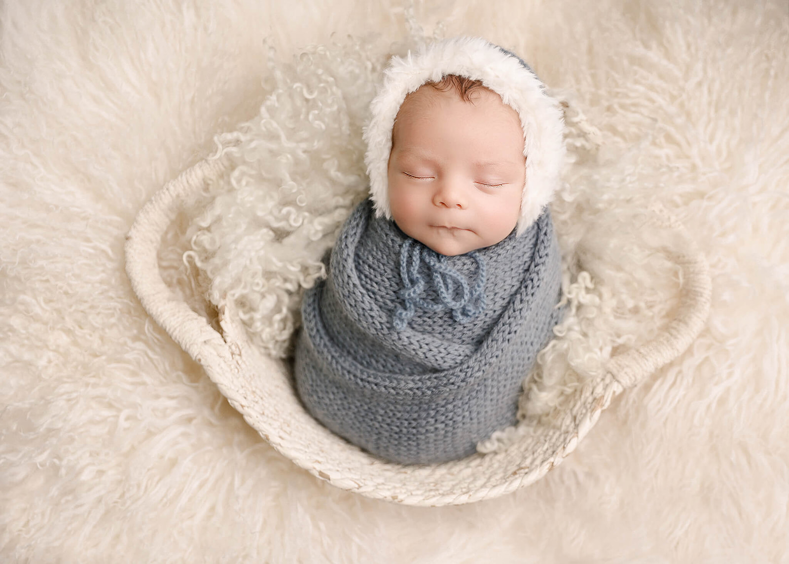 A newborn baby sleeps in a white basket on a fur blanket in a blue knit swaddle