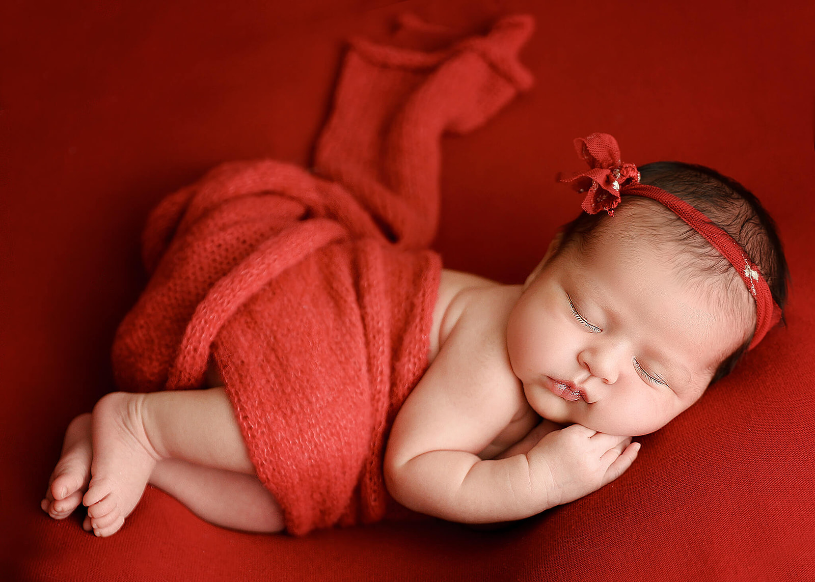 A newborn baby sleeps while wrapped in a red knit blanket wearing a red headband with a bow