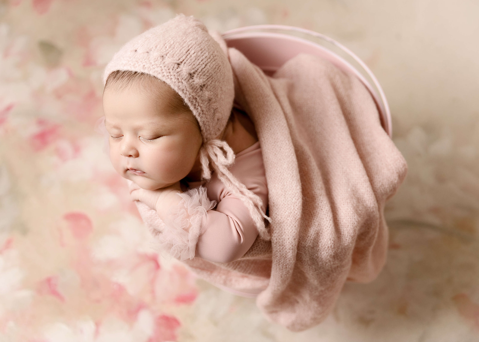 A newborn baby sleeps on her hands in a pink bucket with a blanket and knit hat
