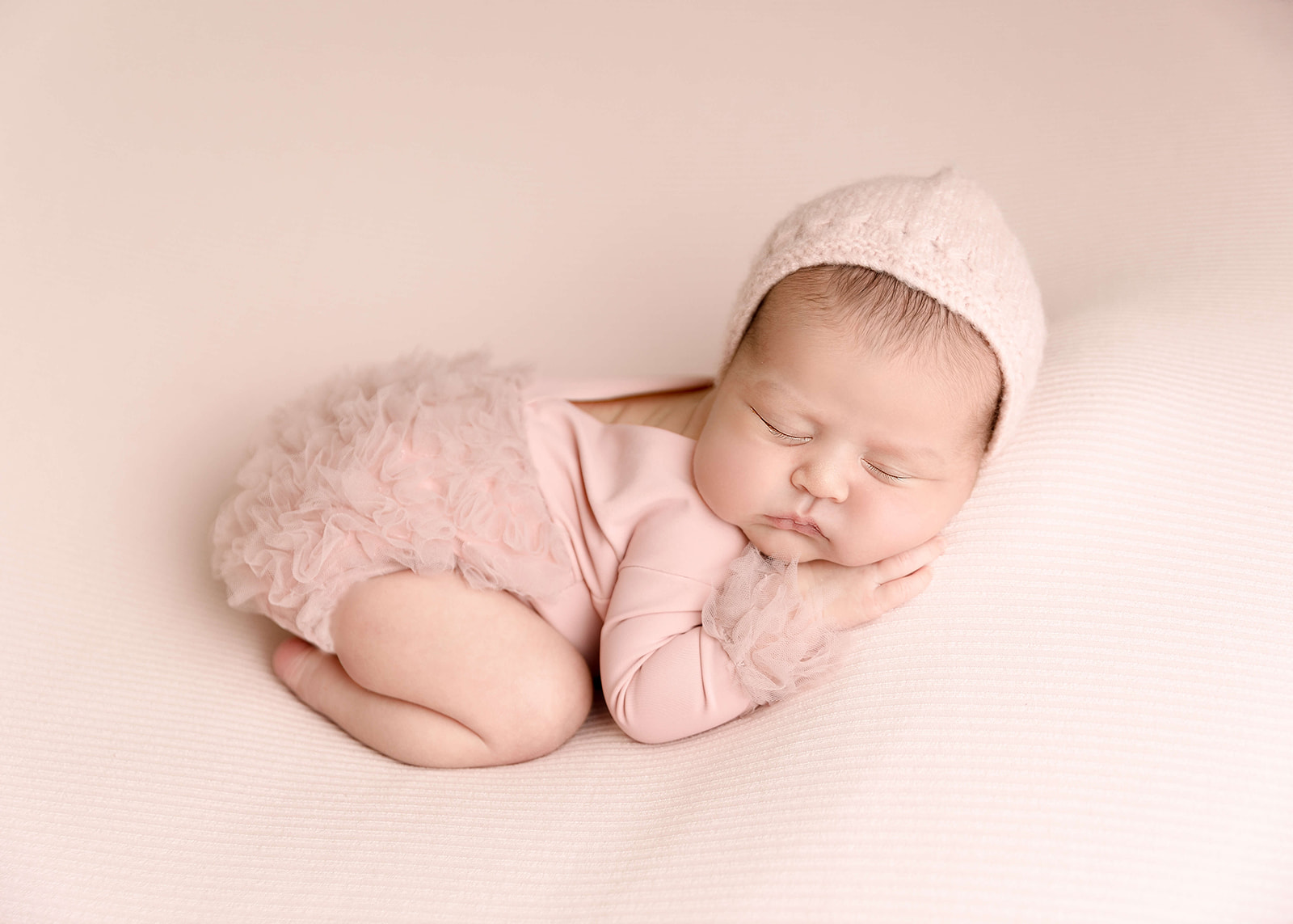 A newborn baby sleeps in froggy pose on a soft pink bed wearing a knit pink hat and pink onesie Los Angeles Midwives