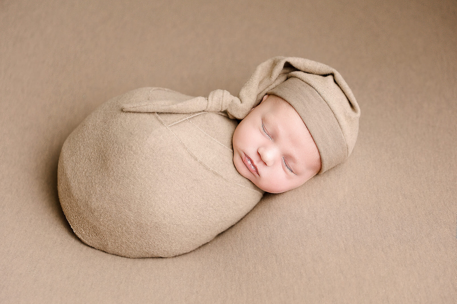 A newborn baby in a brown sleeper cap and swaddle sleeps on a matching bed
