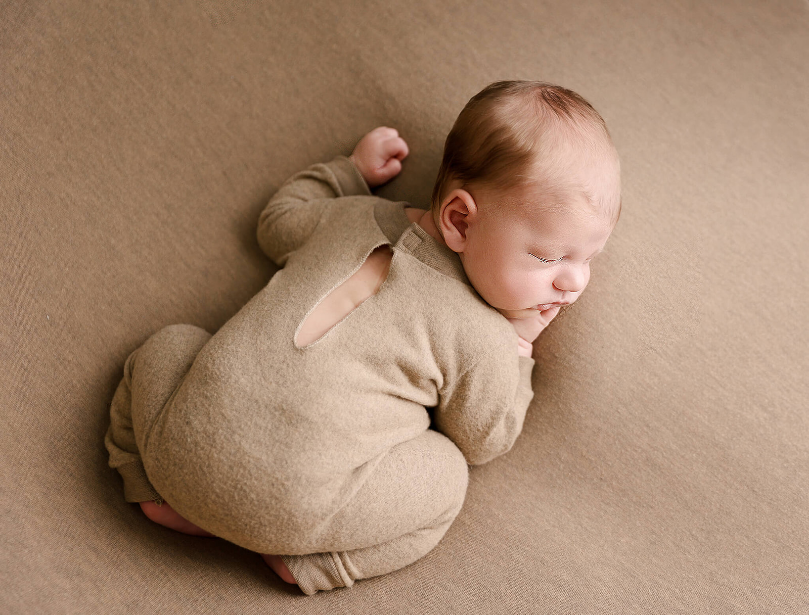 A newborn baby in a brown onesie sleeps in a froggy pose