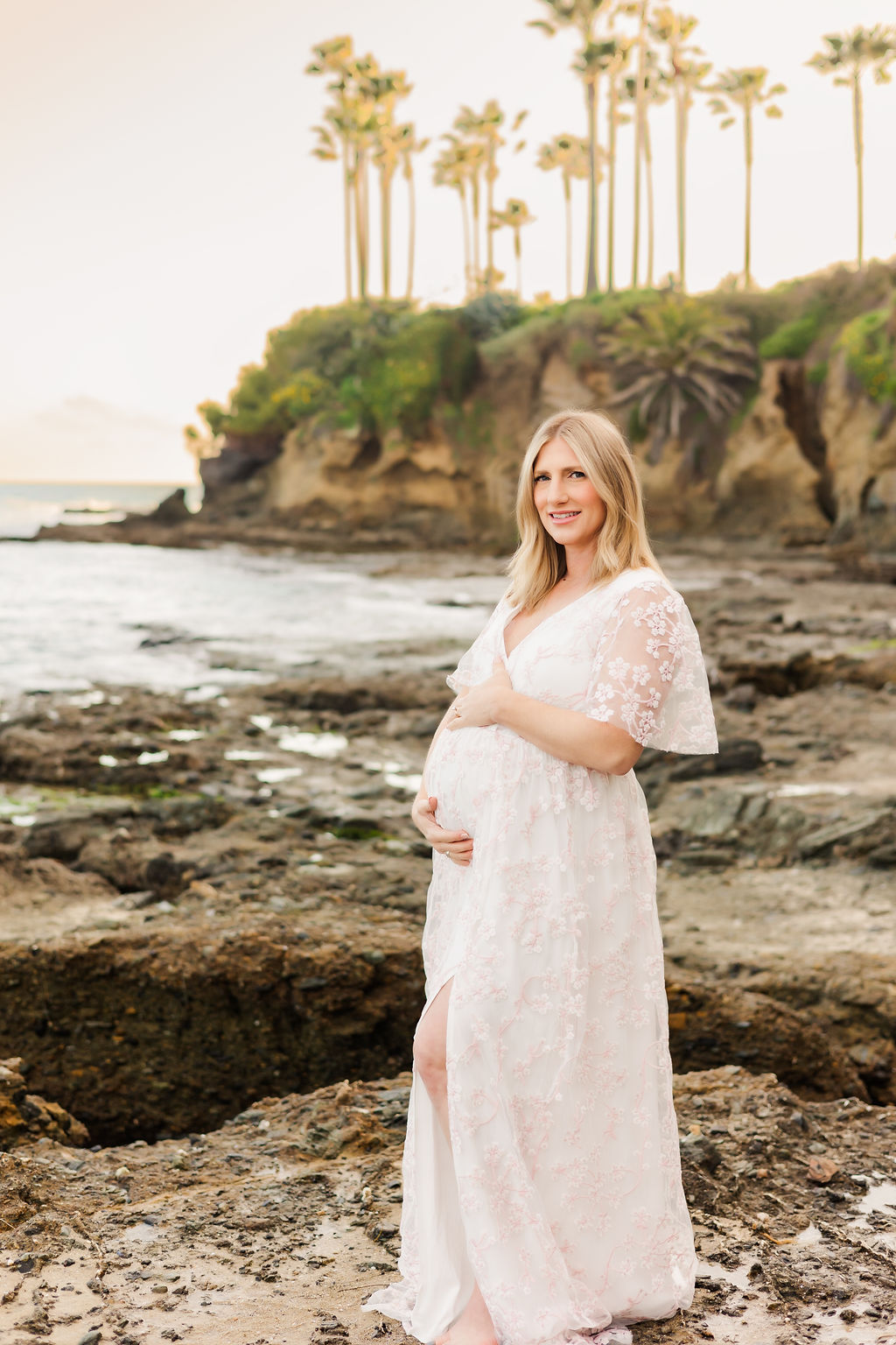 A mother to be in a long white lace maternity gown stands on a rocky shore with a steep cliff behind her
