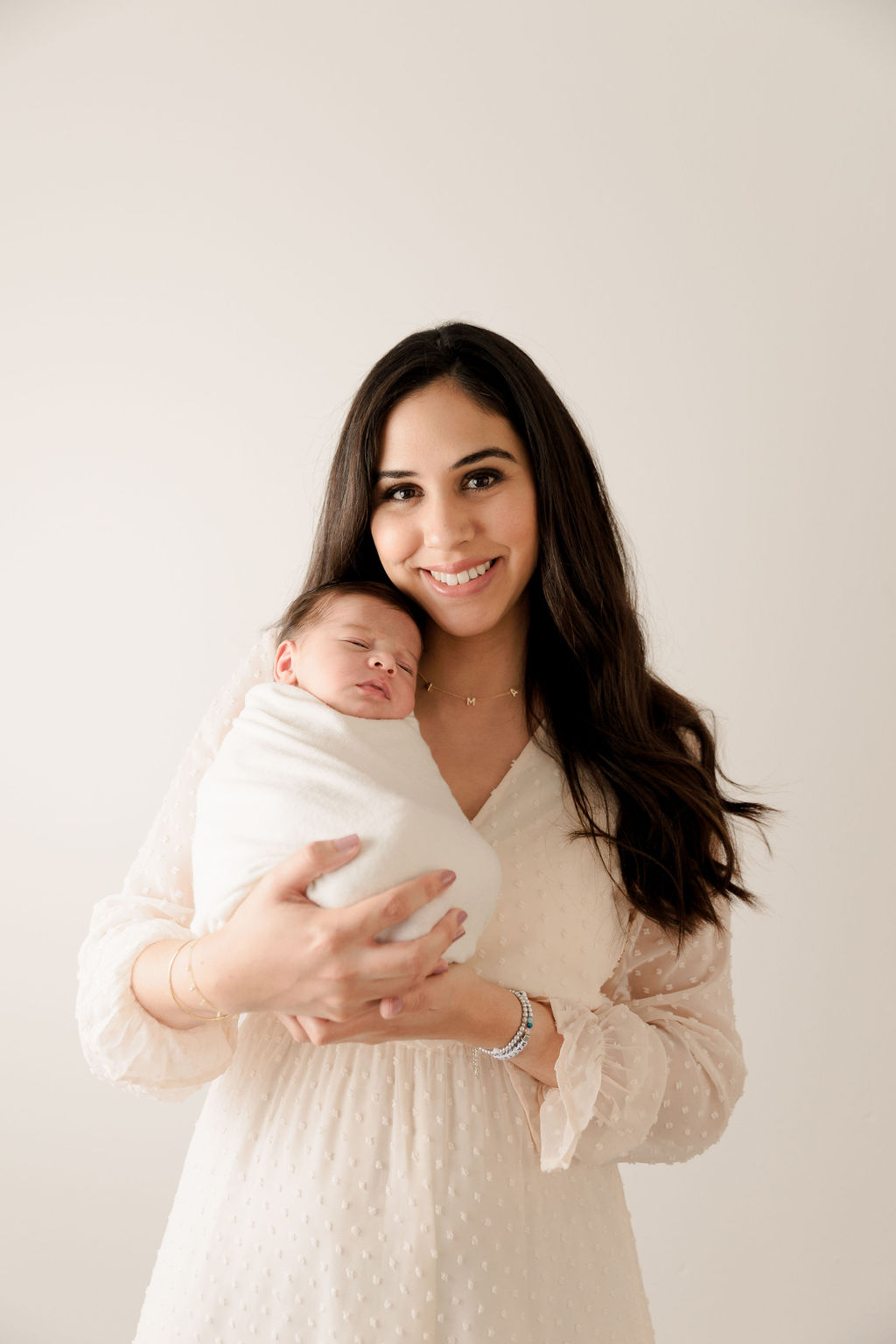 A mother stands in a studio in a white dress holding her sleeping newborn baby