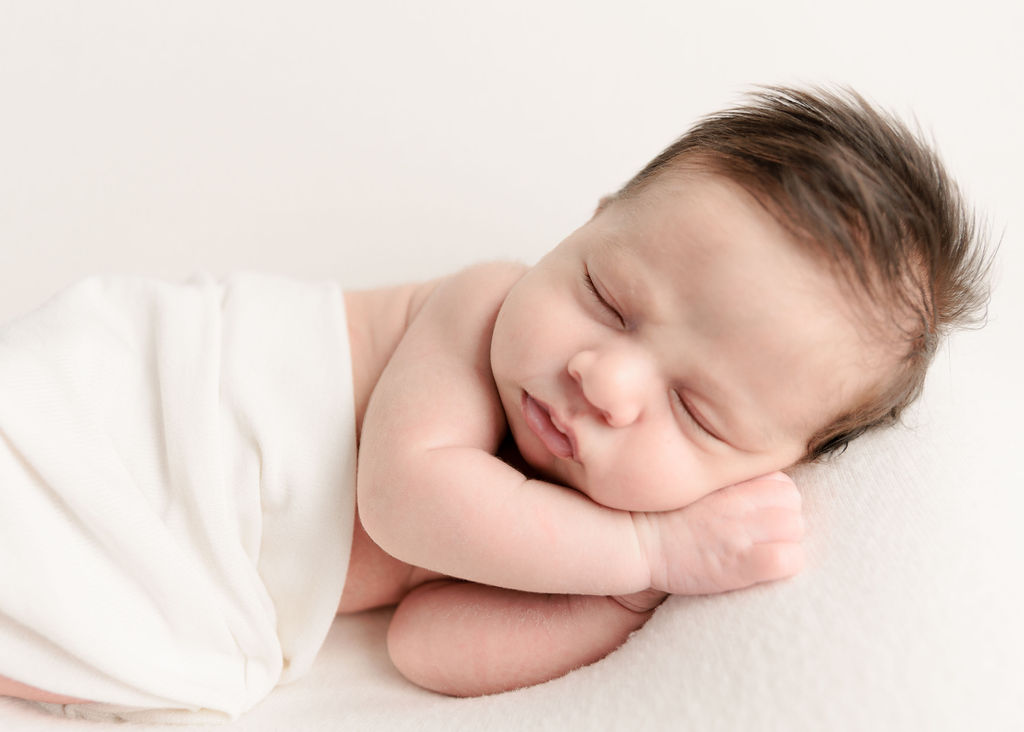 A newborn baby sleeps in its hands under a white blanket neiman marcus newport beach