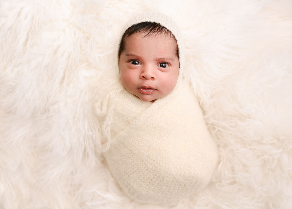A newborn baby lays in a white swaddle with eyes wide open on a furry blanket