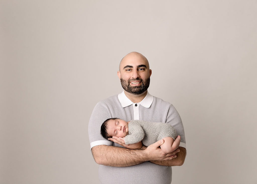 A father in a grey and white polo stands in a studio holding his sleeping newborn baby