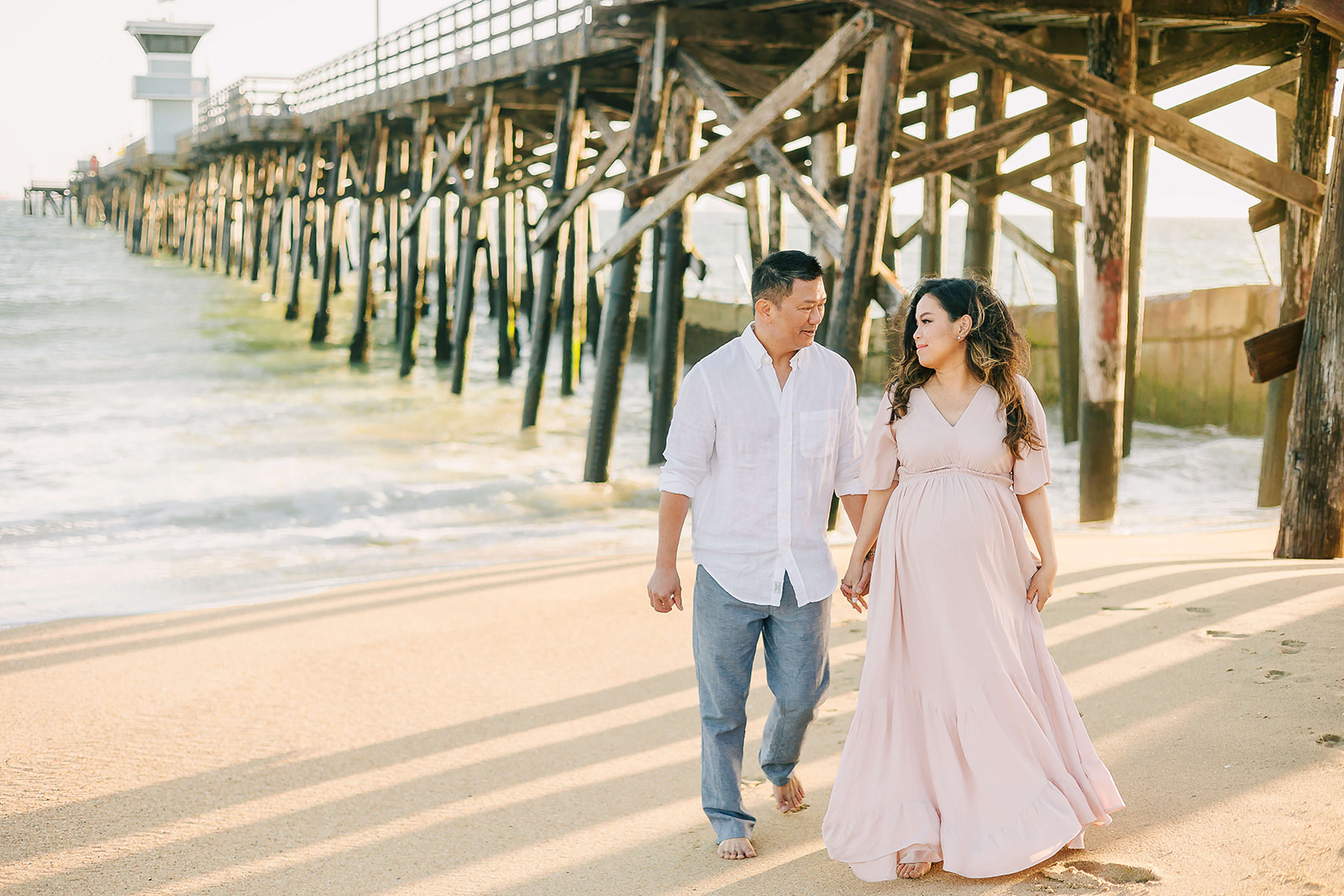 A mother-to-be walks under the pier on a beach while holding hands with her husband