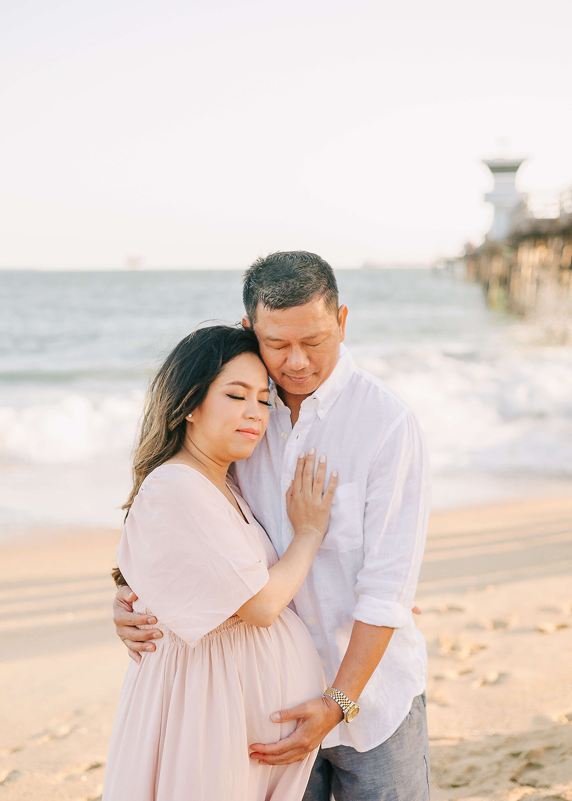 A mother to be cuddles in the chest of her husband while standing on the beach by a pier Best Maternity Hospital in Los Angeles
