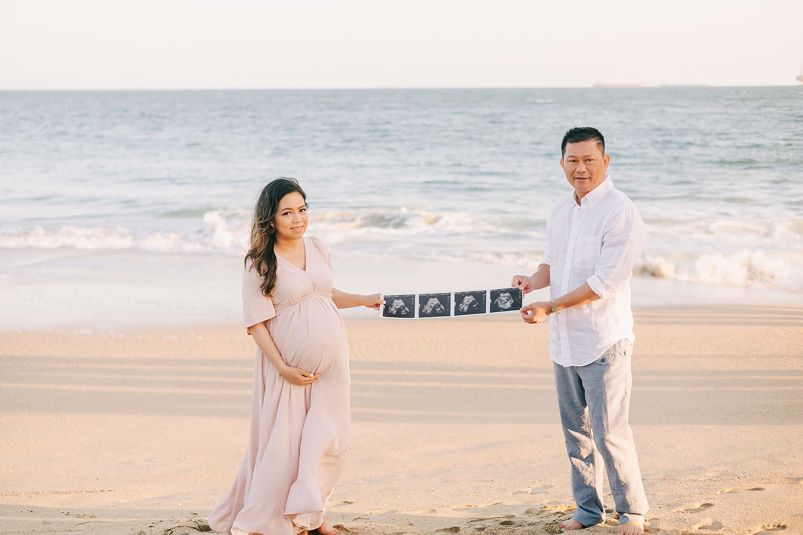 A mother to be and father stand on a beach holding their sonogram strip between them