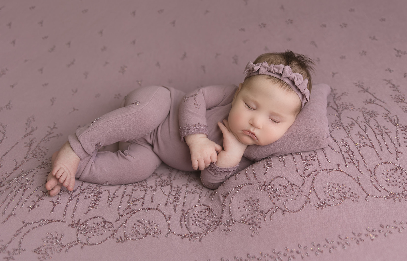 A newborn baby sleeps on her side in a purple onesie and matching headband