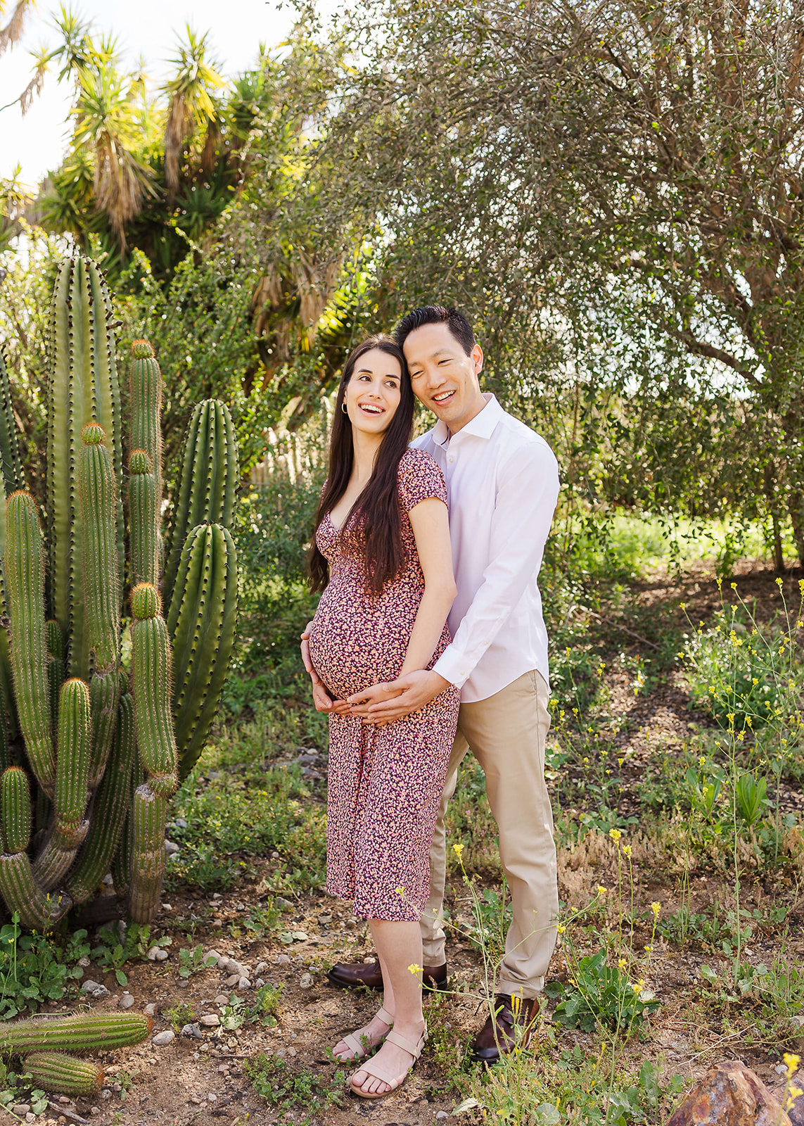 A mother to be holds her bump in a purple dress while standing in a desert trail by a tree and cactus