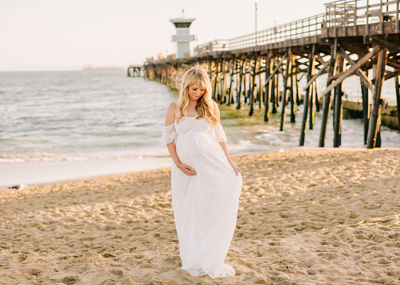 A mother to be walks down a beach playing with her white maternity gown by the pier