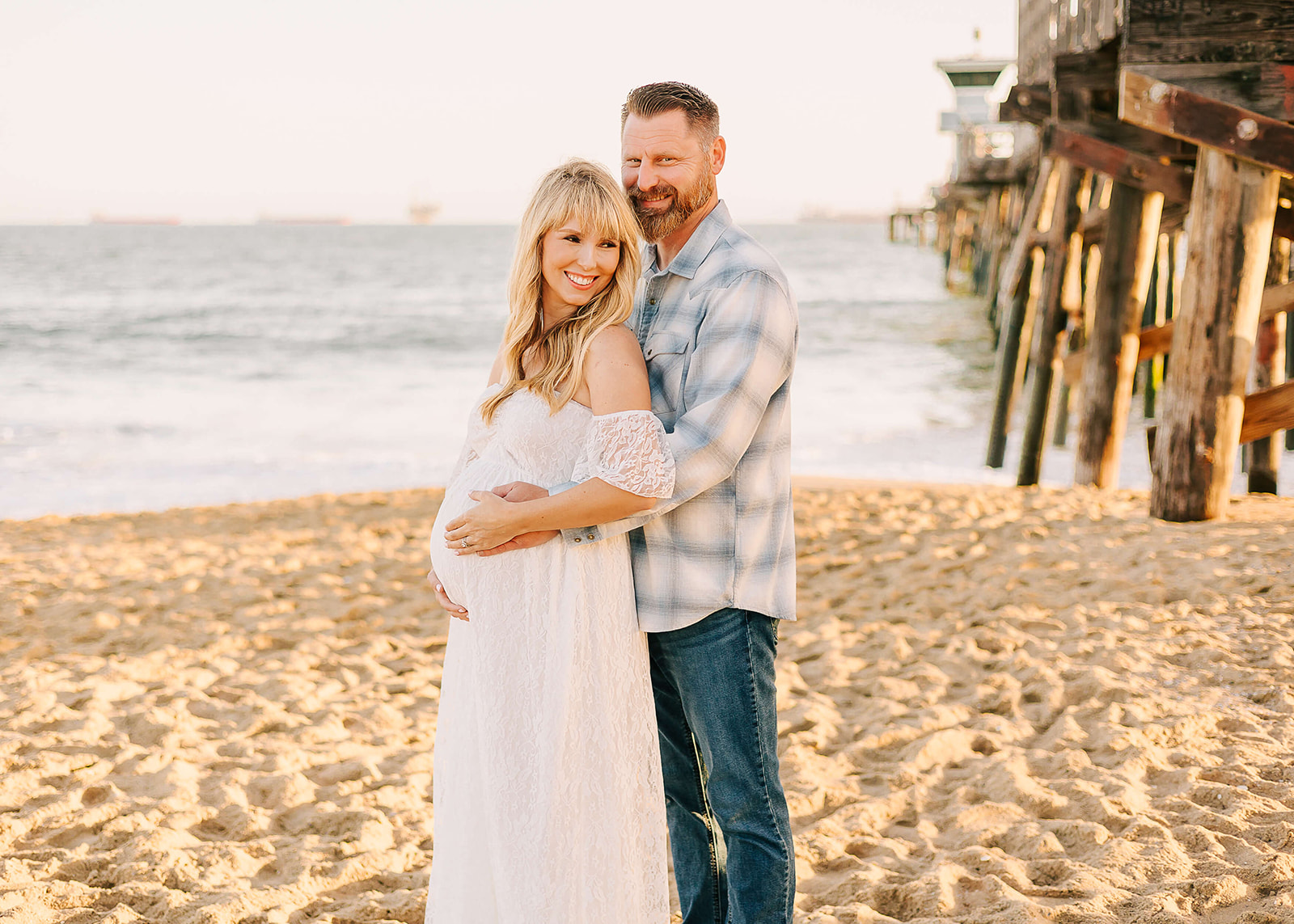A mother to bee stands on the beach next to the pier while her husband hugs her and the bump from behind