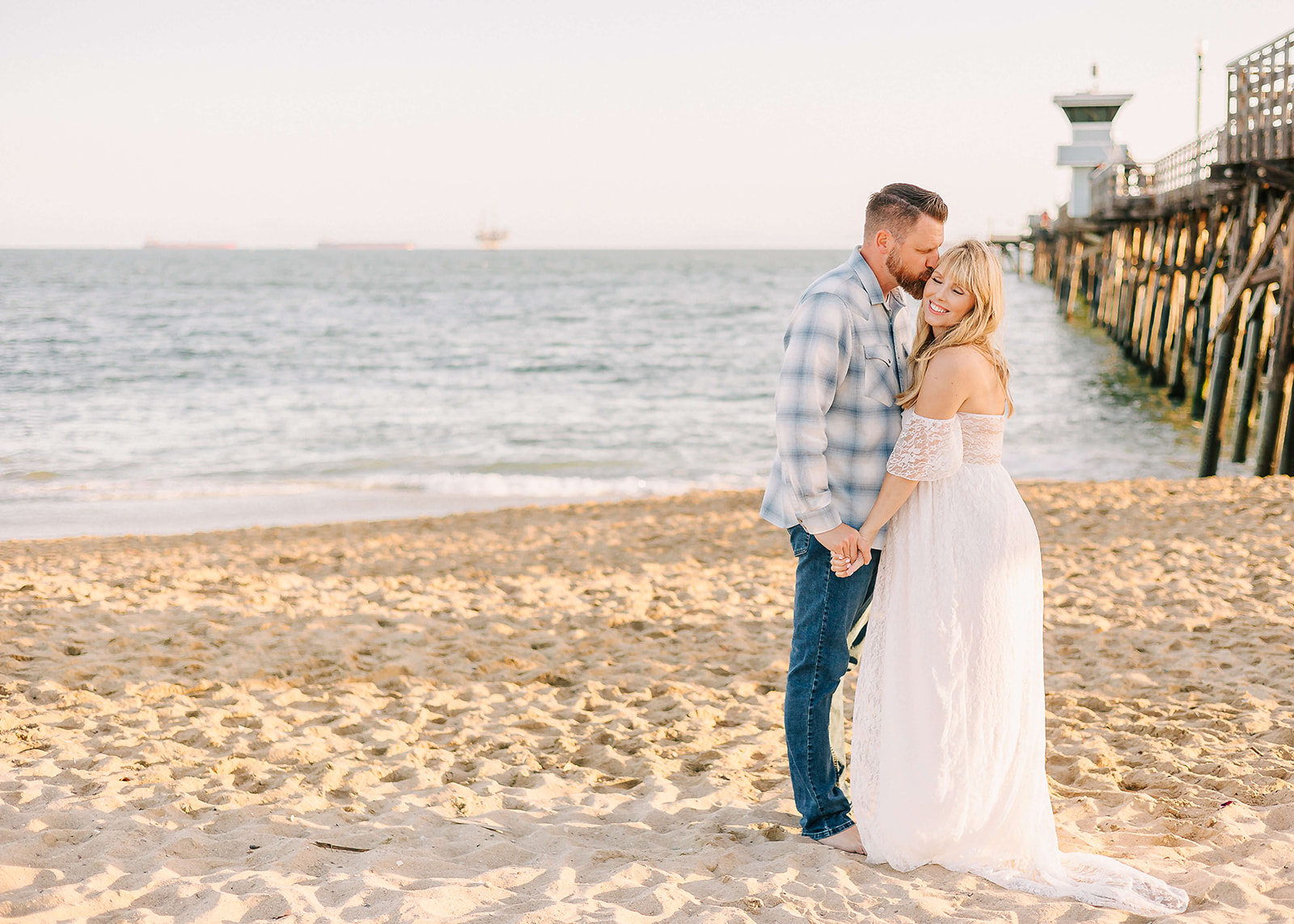 A husband kisses the temple of his pregnant wife on the beach wearing a white lace maternity gown Los Angeles Pediatricians