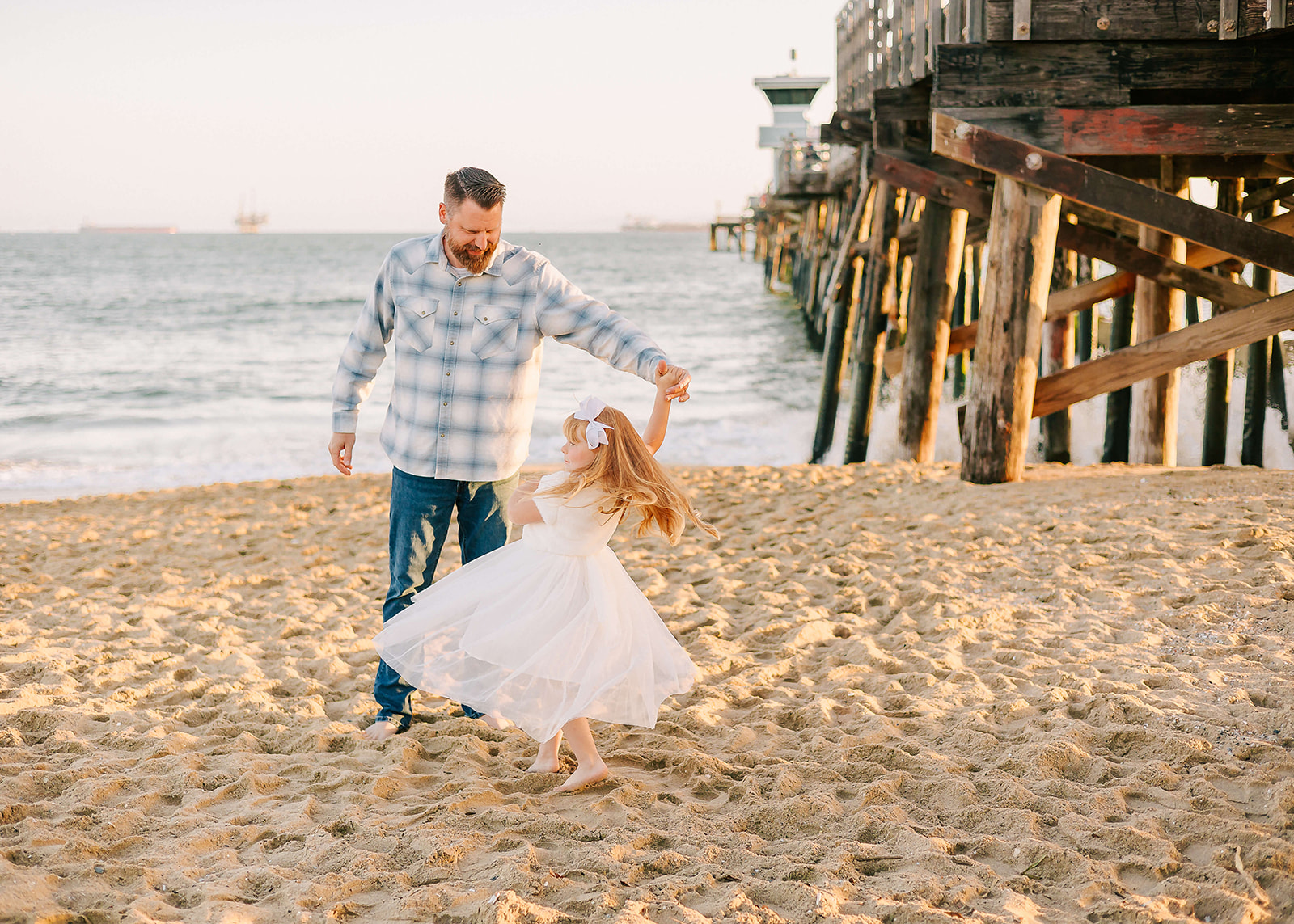 A father in blue flannel dances with his young daughter in a white dress under the pier on the beach Los Angeles Pediatricians