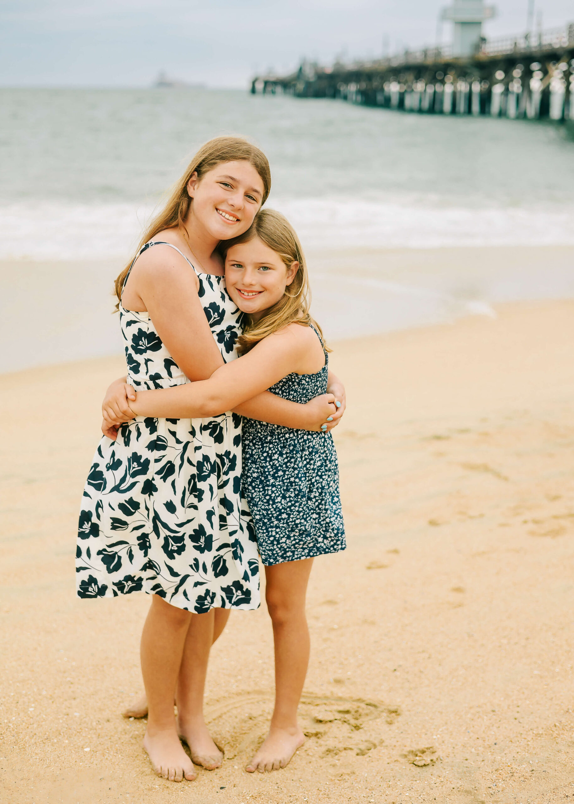 Sisters hugging on beach during family session smiling.