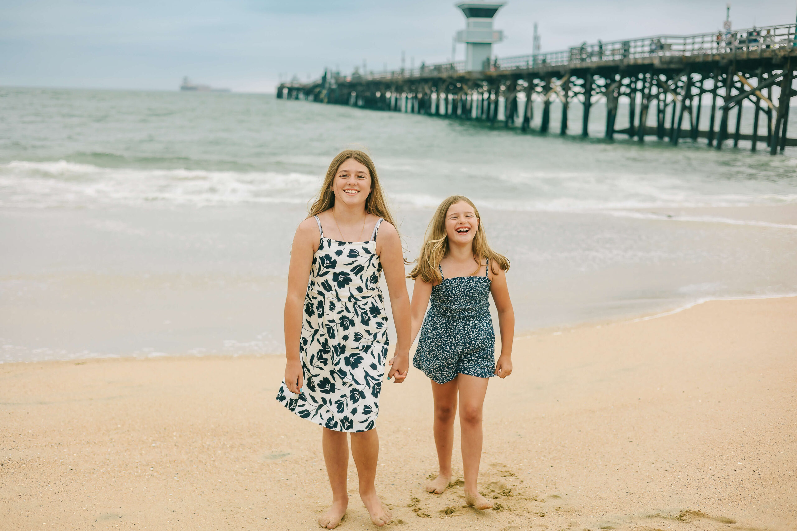 Sisters holding hands laughing while walking on beach during session by Ashley Nicole.