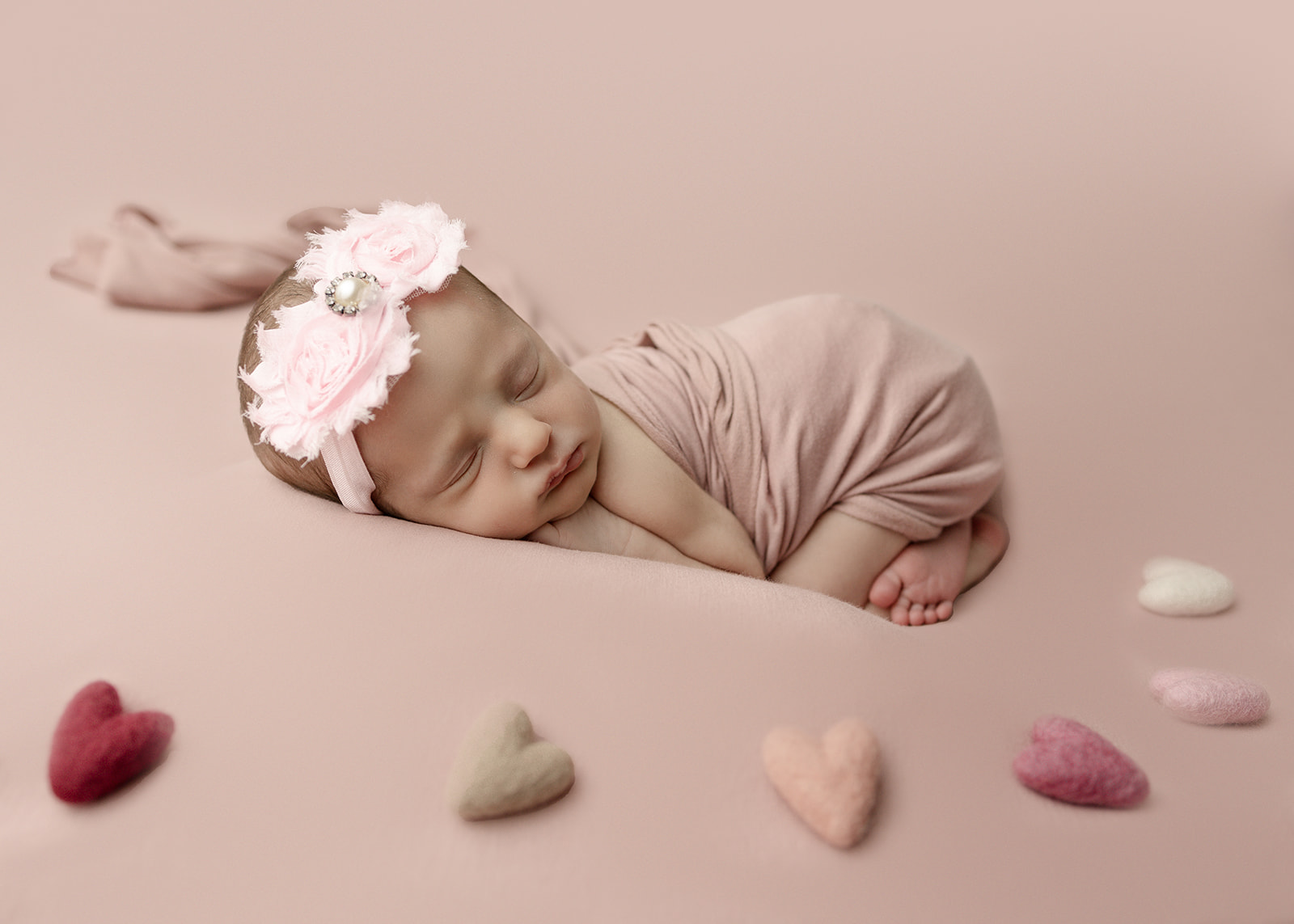 A newborn baby sleeps in a pink headband and swaddle while surrounded by felt hearts Huntington Beach Pediatricians