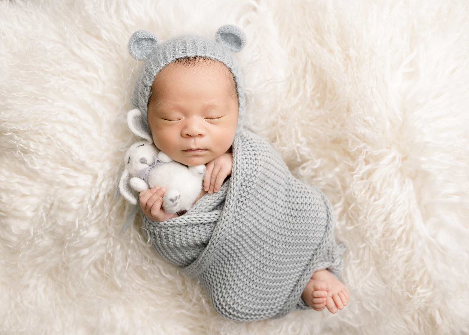 A newborn baby sleeps while cuddling a stuffed bunny in a knit swaddle and bonnet with ears Irvine Midwife