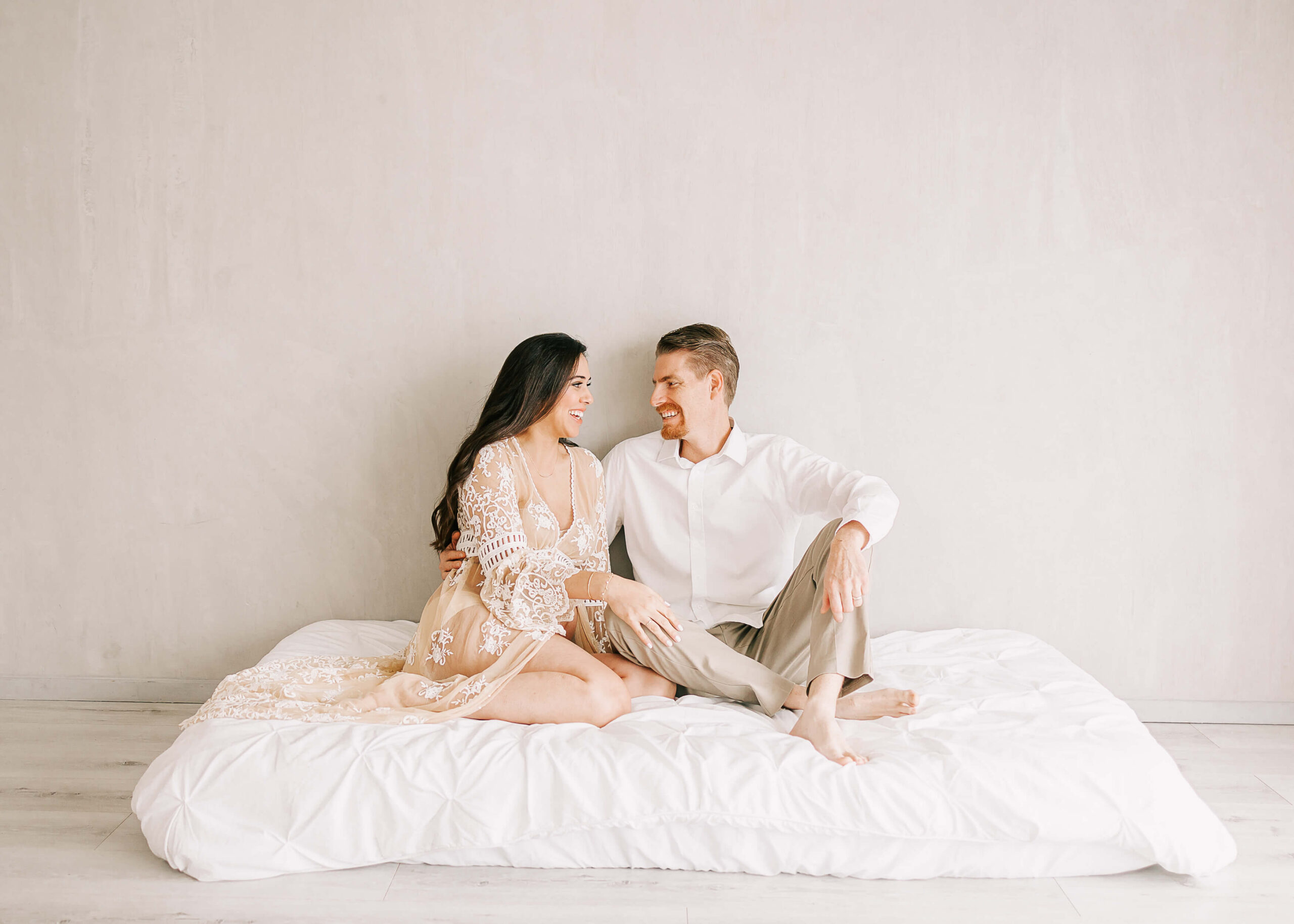 Excited expectant parents laughing and chatting on bed in studio by Ashley Nicole Photography.