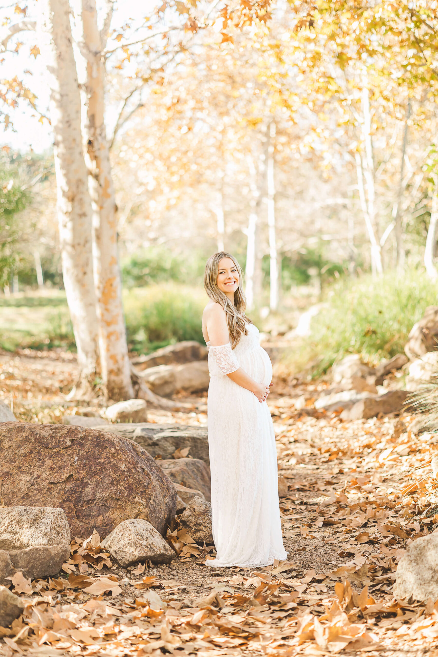 Mama posing holding her baby bump on a nature trail wearing a white lace dress.