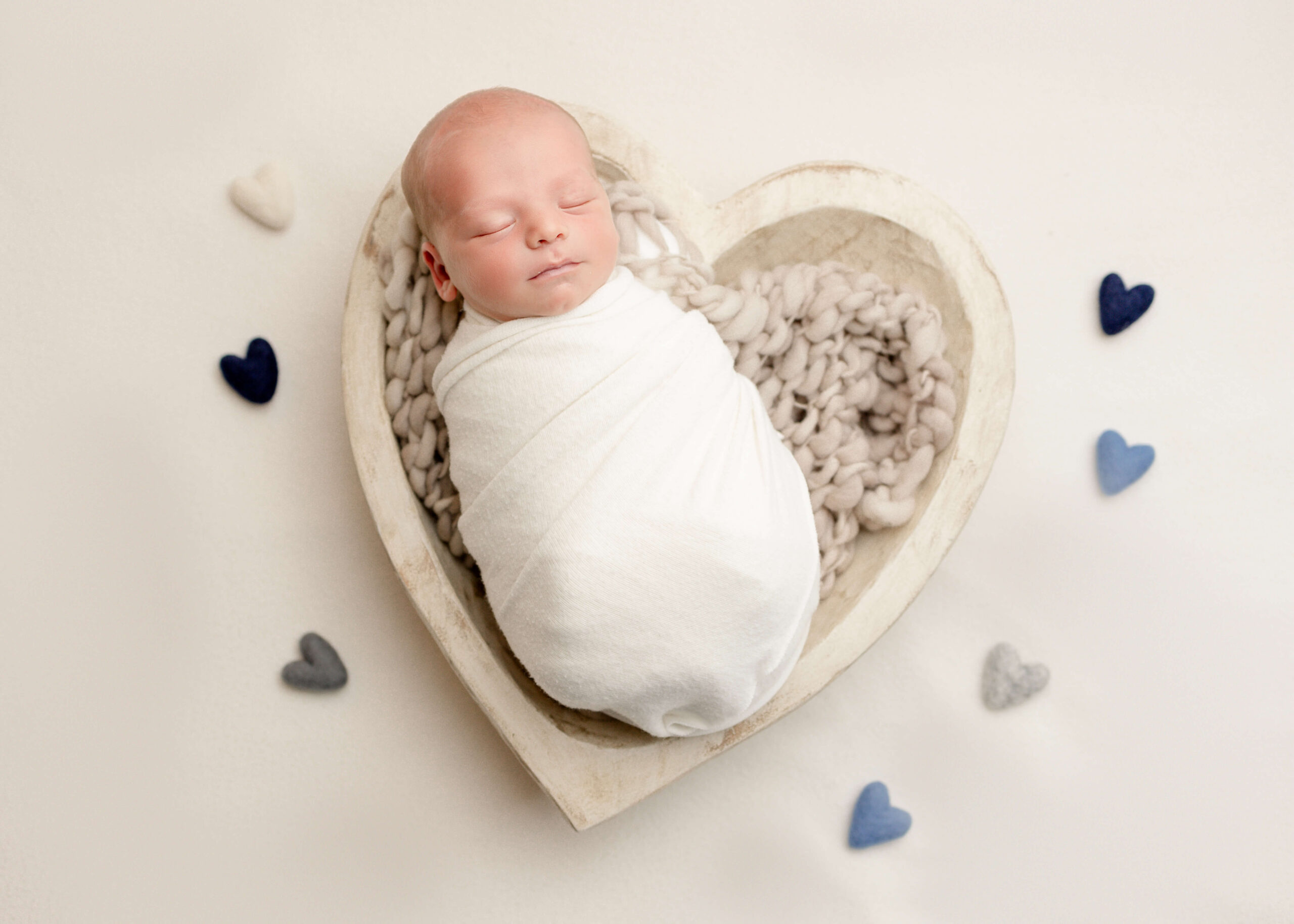 Baby boy wrapped in cream placed in white heart bowl with felt hearts placed around him.
