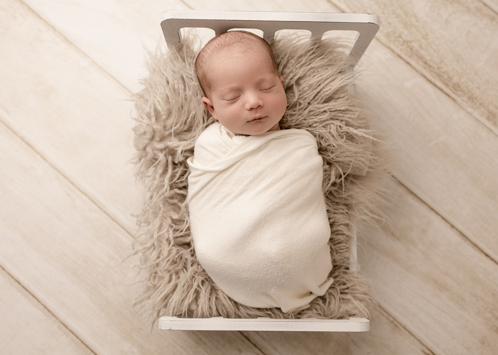 A newborn baby sleeps in a white swaddle on a fur blanket in a tiny bed