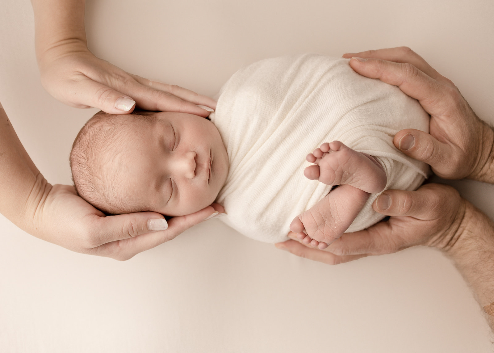 A newborn baby in a white swaddle with feet sticking out is held by mom and dad's hands