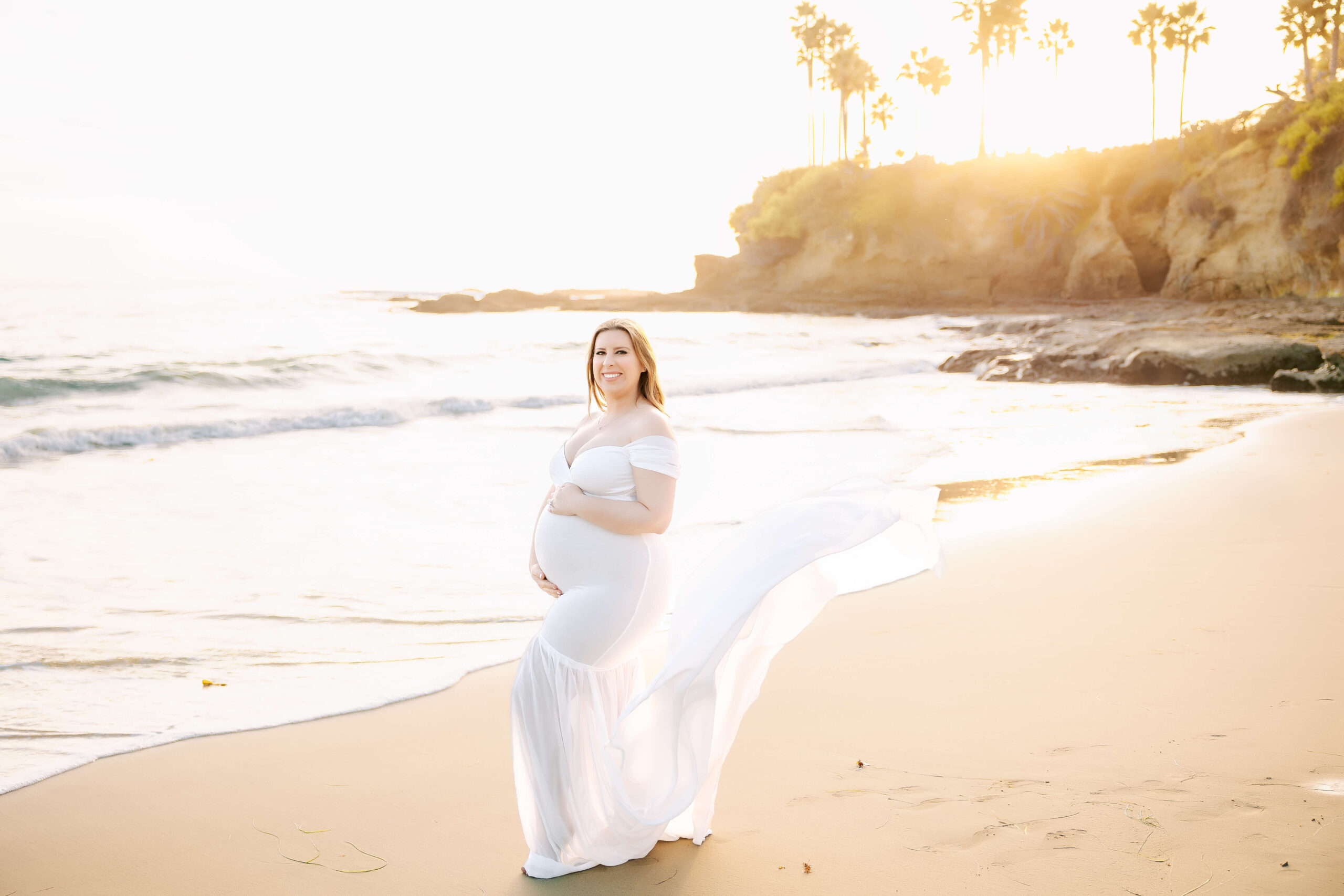 Expectant Mama posed in maternity session holding belly while dress train flows in the breeze in Laguna Beach by Ashley Nicole Photography.