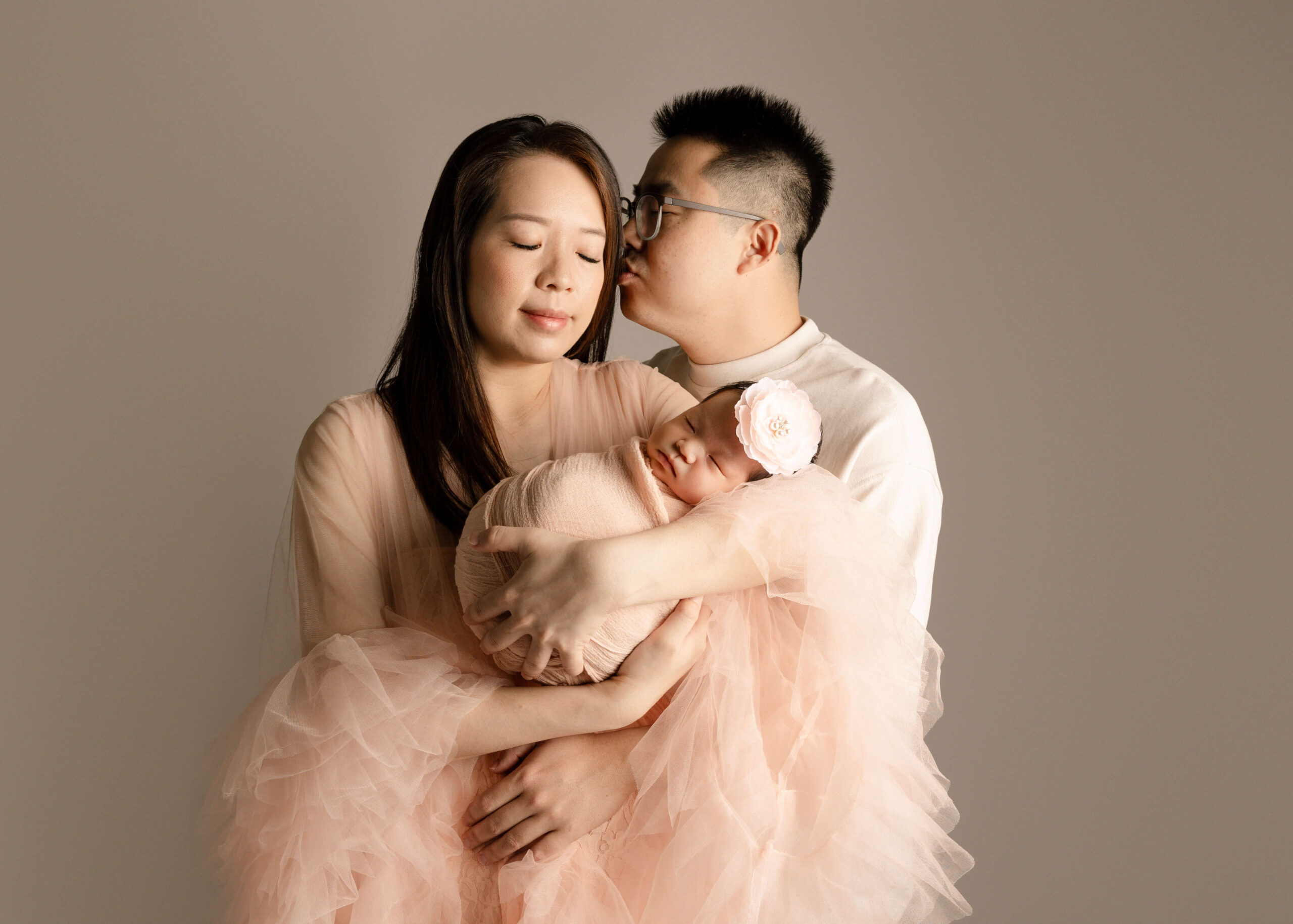 Husband kissing side of wife's face while they are embraced, and wife is holding their new baby girl. In studio in Los Angeles by Ashley Nicole.