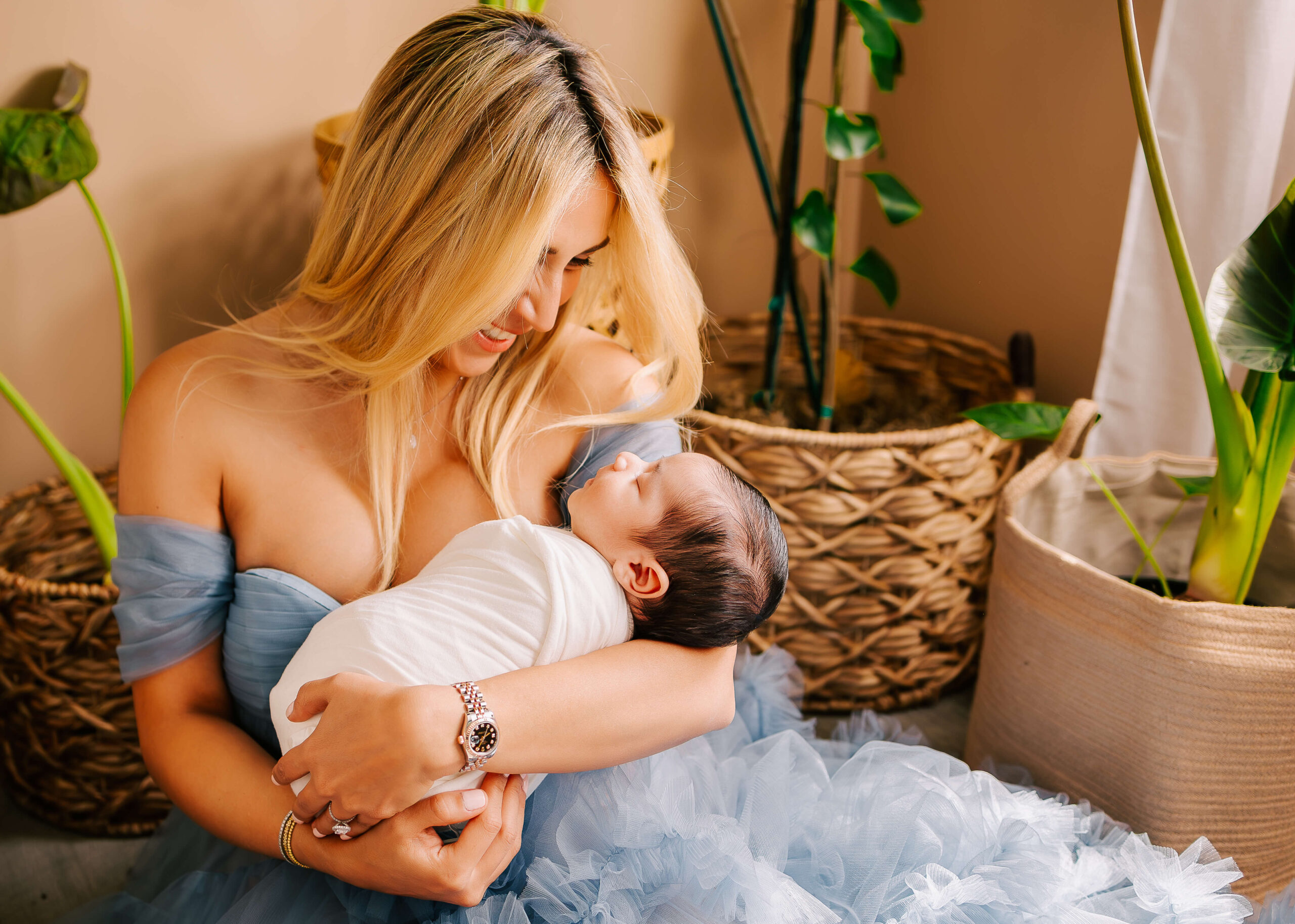Mom sitting on floor next to bed snuggling her baby during newborn lifestyle session in Orange County CA studio by Ashley Nicole.