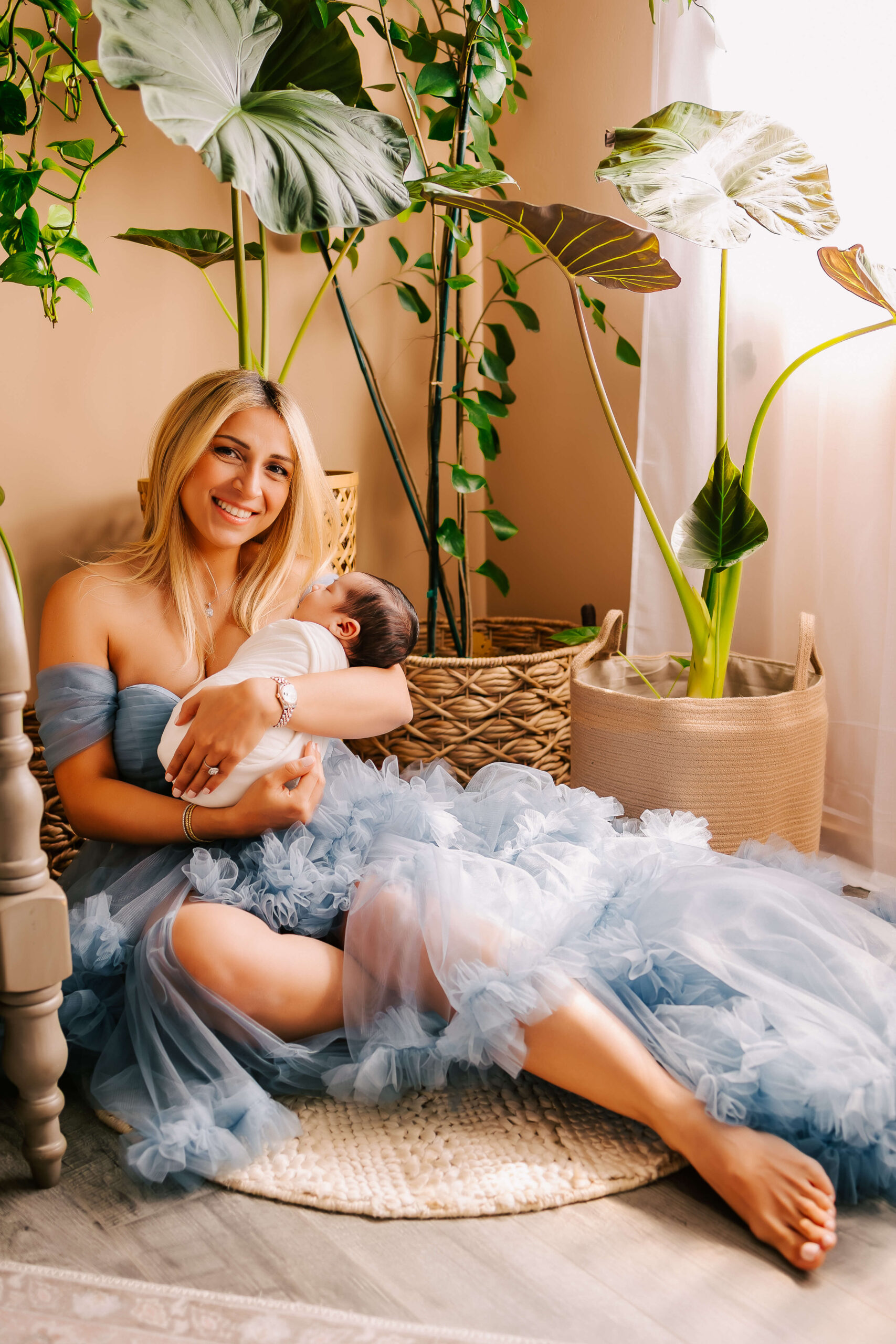 Mom sitting on floor next to bed holding her baby and smiling looking at camera during newborn lifestyle session in Orange County CA studio by Ashley Nicole Photography.