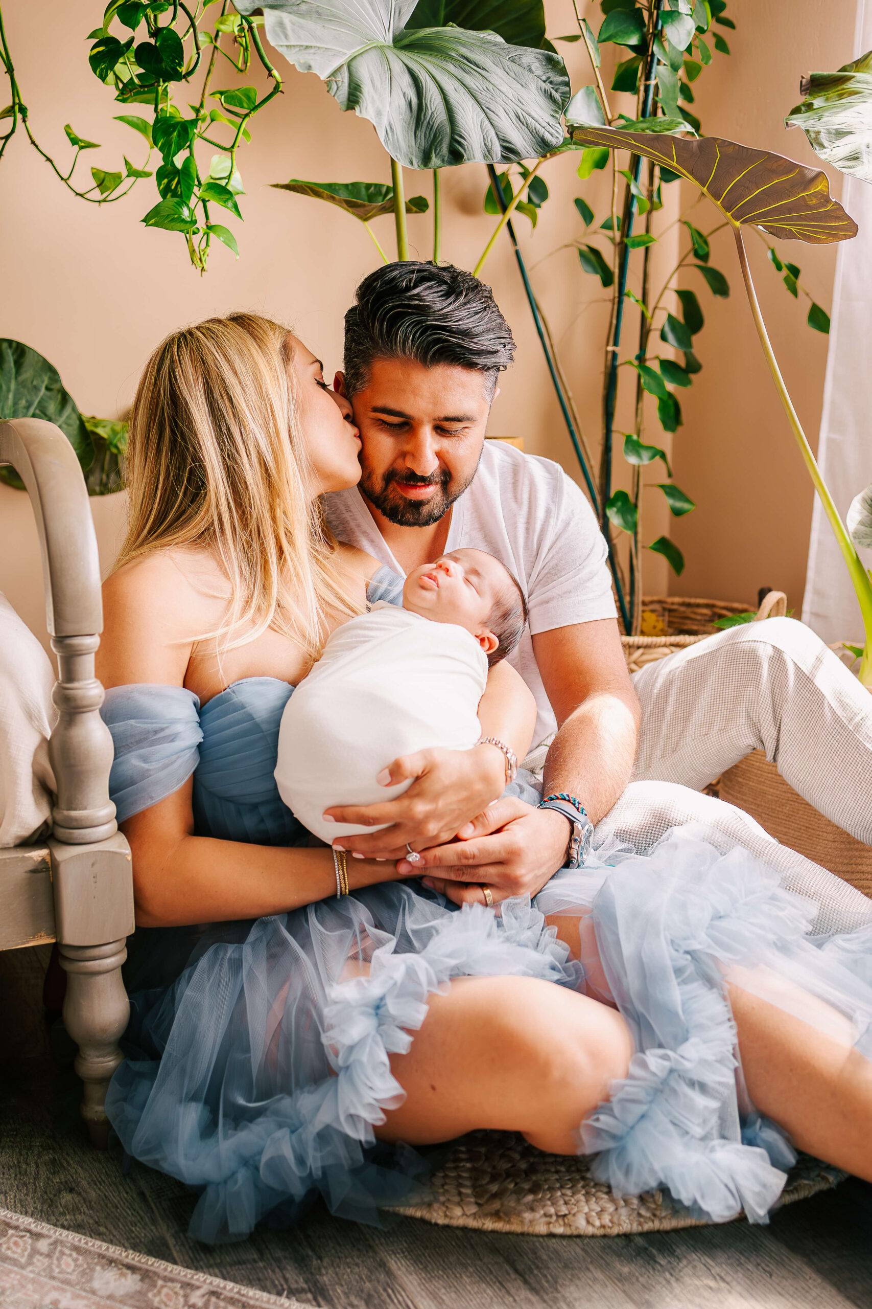 Mom and dad sitting on floor next to bed snuggling their baby and mom is kissing the dad on the cheek, during newborn lifestyle session in Huntington Beach, CA studio by Ashley Nicole.