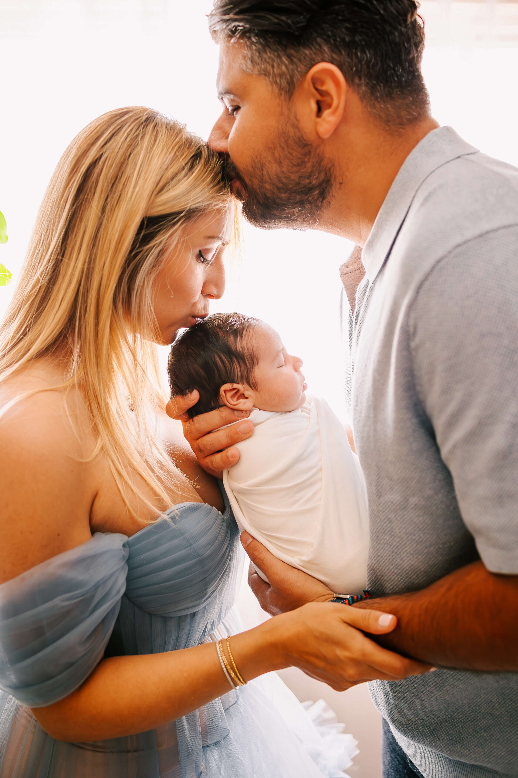 Dad is kissing moms forehead while mom kisses the top of babies head in front of big window in Orange County Studio by Ashley Nicole. For Postpartum Doula Los Angeles.