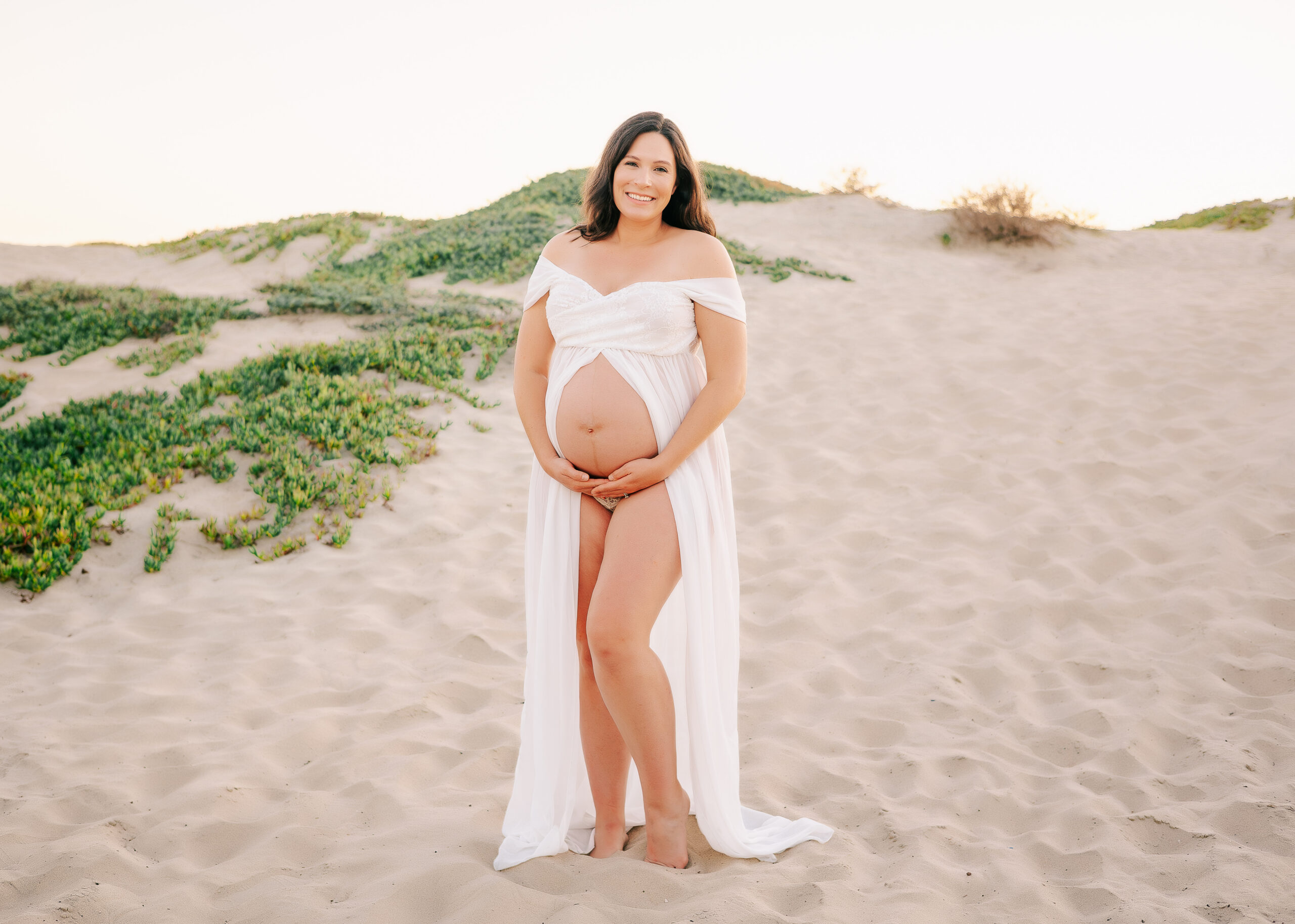 Pregnant Mama wearing white open belly dress standing in sand dunes in Seal Beach by Ashley Nicole.
