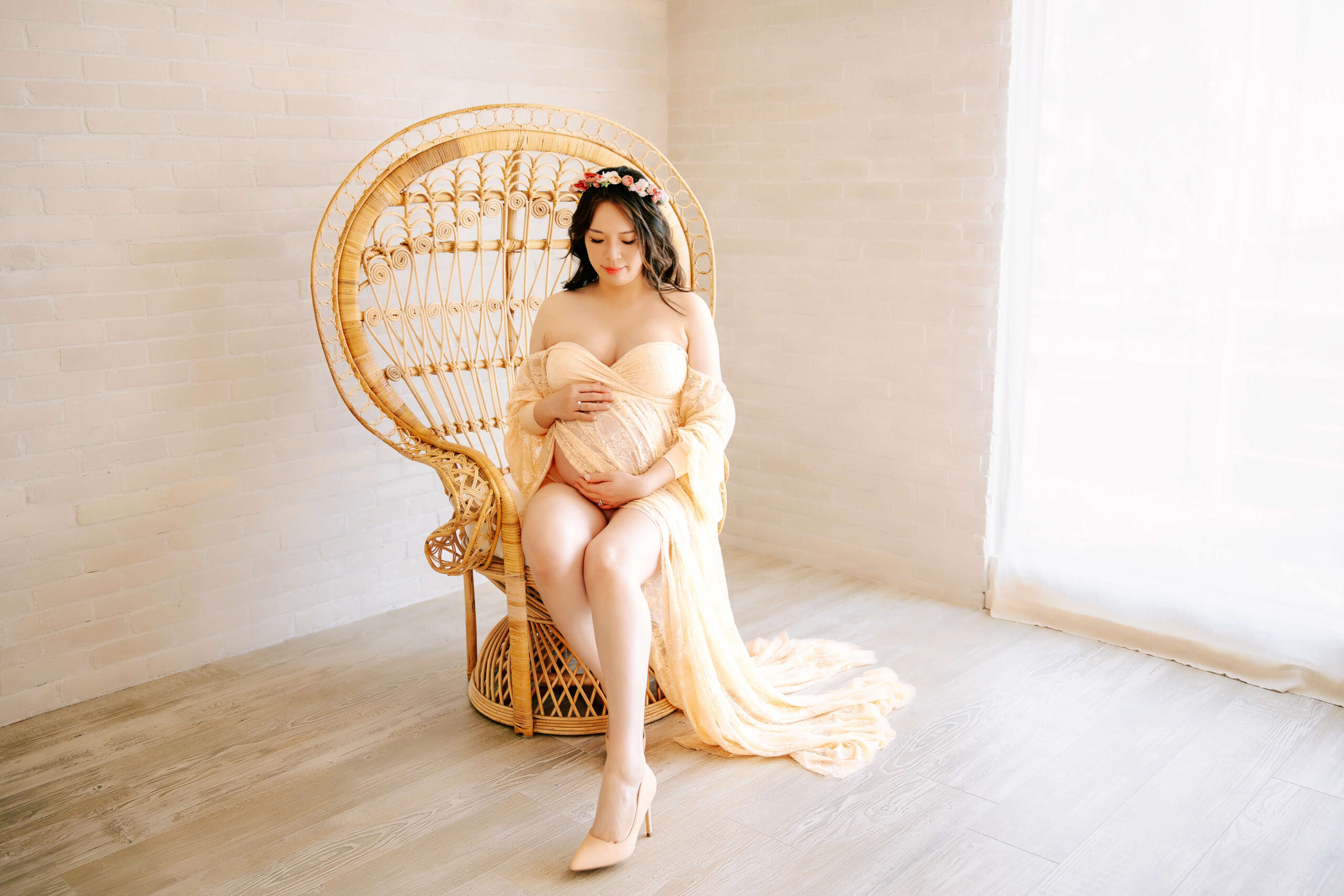 Beautiful expectant mama wearing a lace dress sitting in Peacock Boho chair looking down at her belly in studio in Orange County, CA by Ashley Nicole.