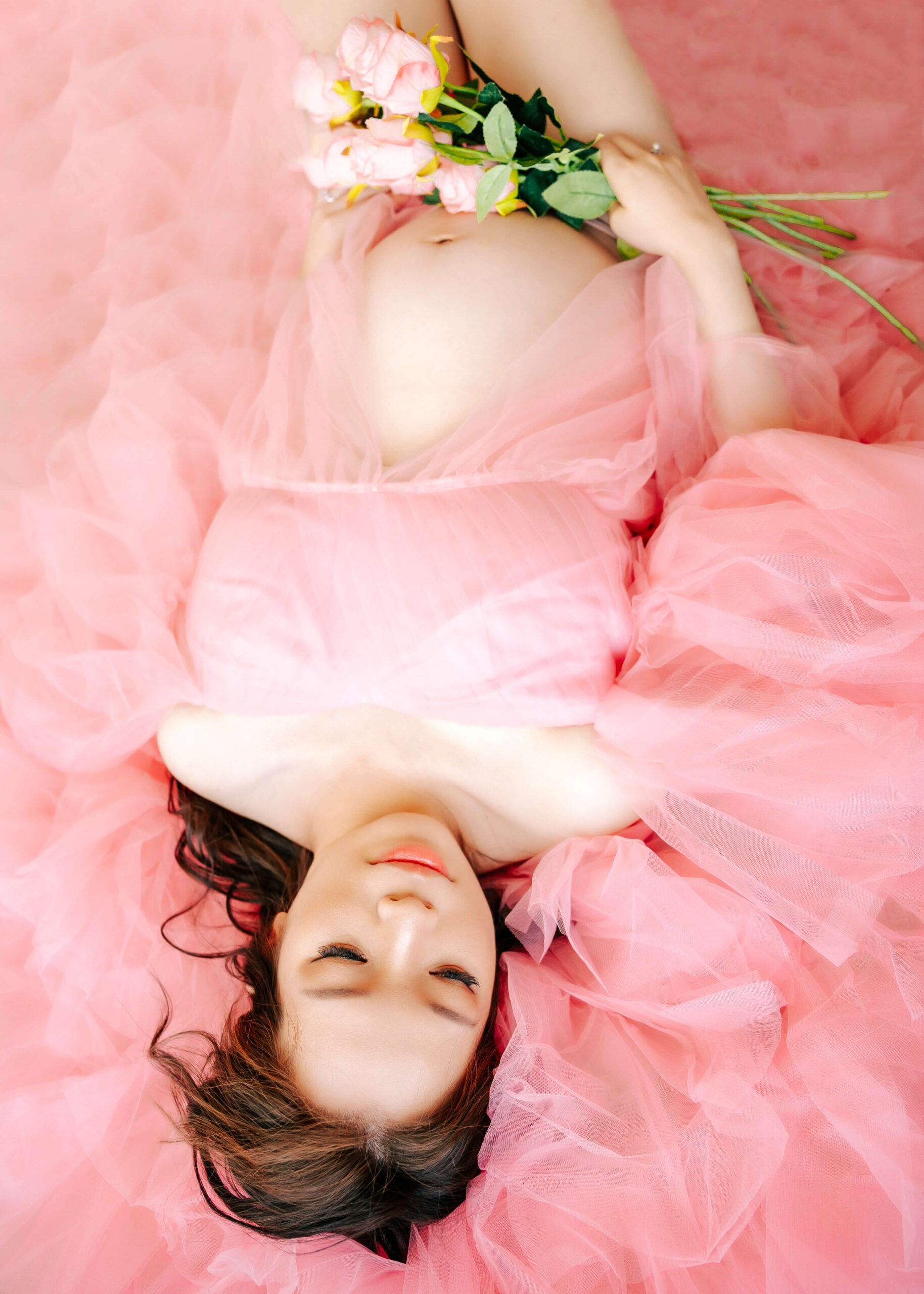 Expectant Mama laying on floor surrounded by beautiful pink tulle dress holding flowers dreaming of her baby in Orange County, CA studio by Ashley Nicole.