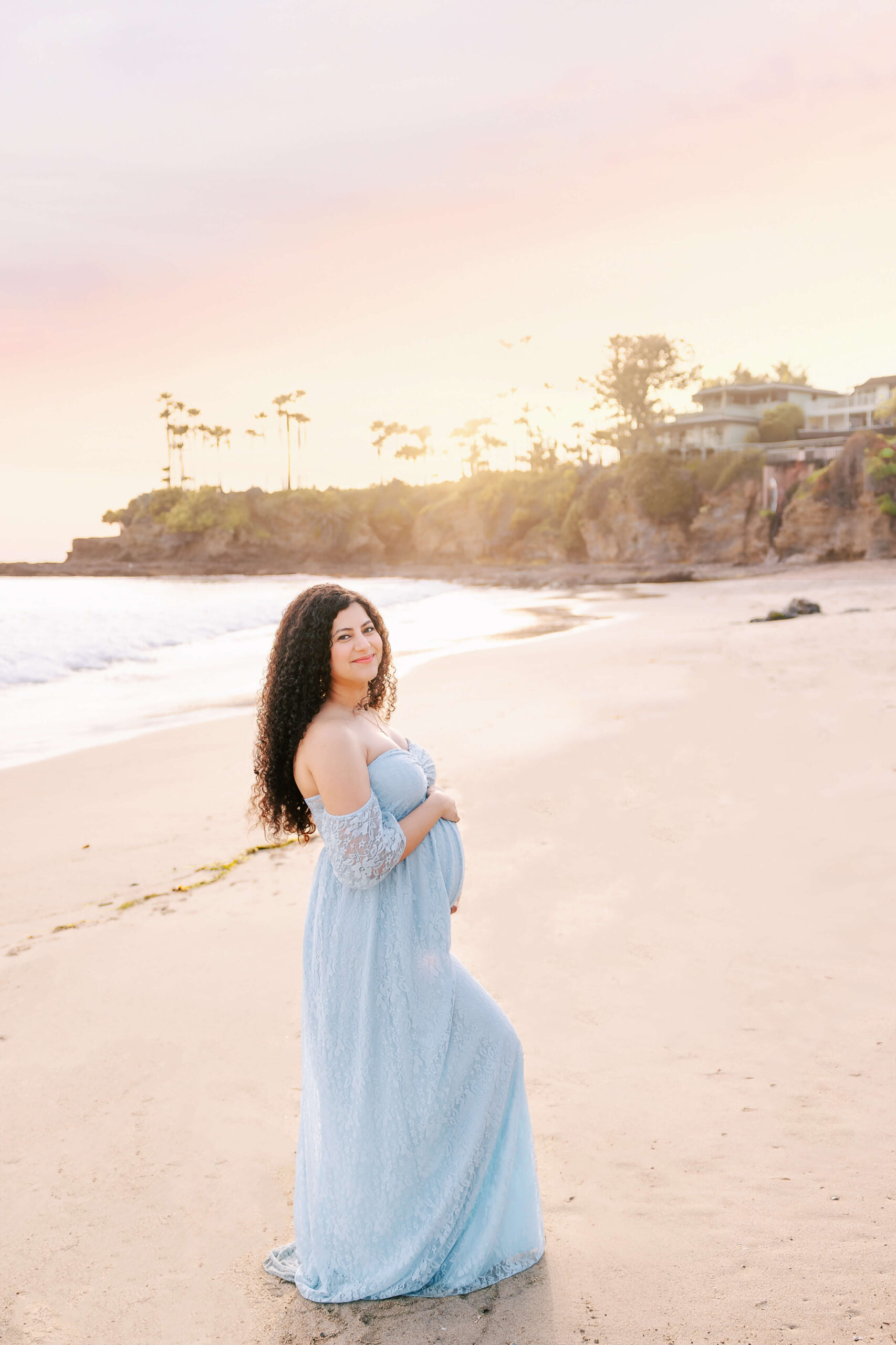 Expectant Mama wearing a blue lace dress holding her bump and smiling in Laguna Beach, CA maternity session by Ashley Nicole Photography.