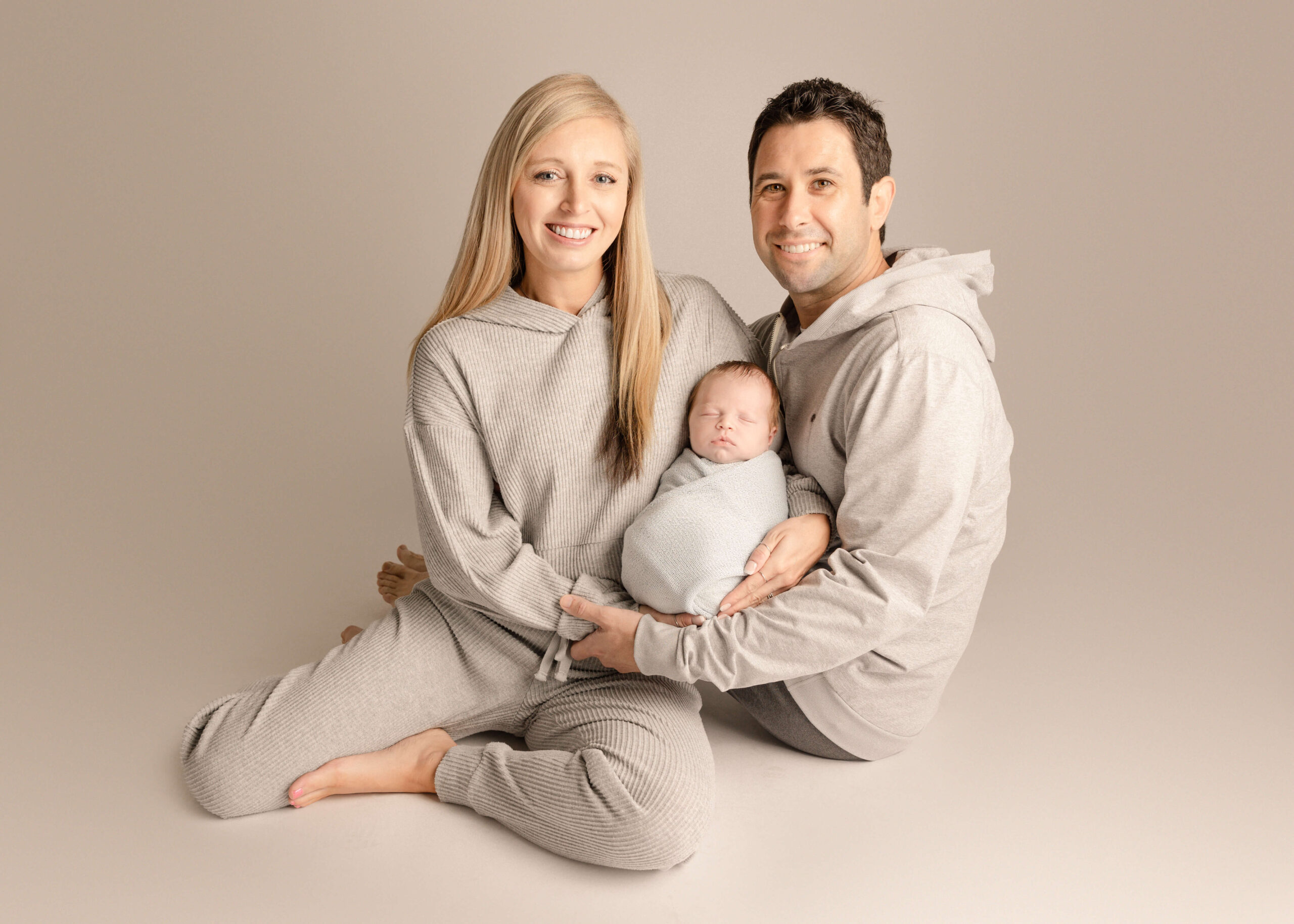 Parents sitting in sweat outfits on floor smiling holding the baby in studio in Orange County, CA by Ashley Nicole Photography.