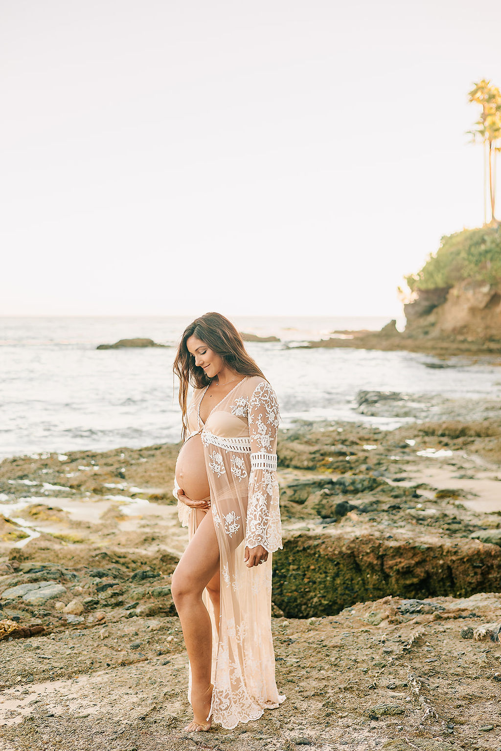 A mom to be in a sheer maternity coverup smiles down at her bump while walking on a rocky beach after her 3D ultrasound in Long Beach