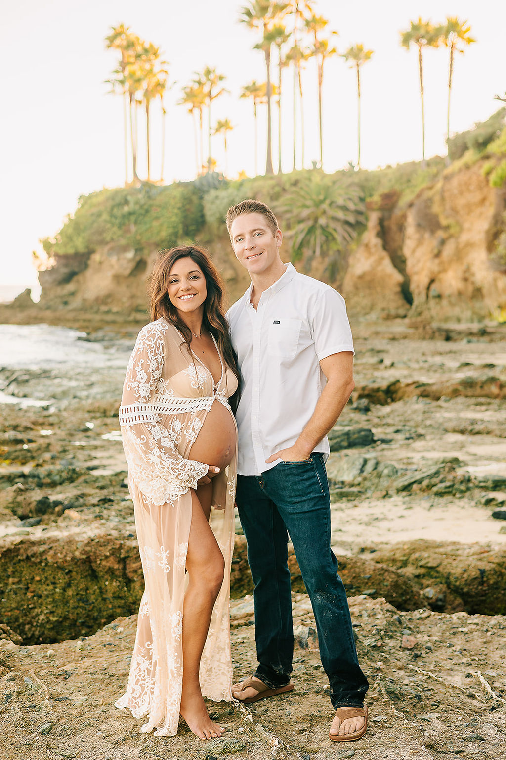 Happy expecting parents stand on a beach together smiling with arms around each other