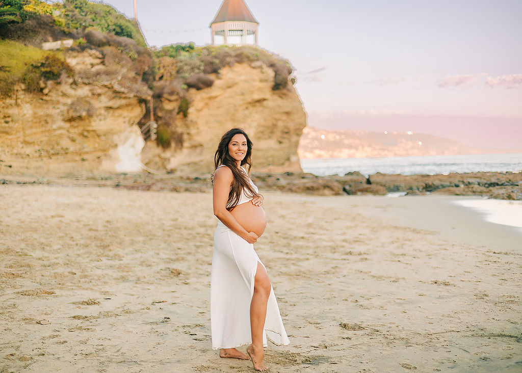 A happy pregnant woman stands on a beach in an open white maternity dress with bump out at sunset after her 3D ultrasound in Long Beach