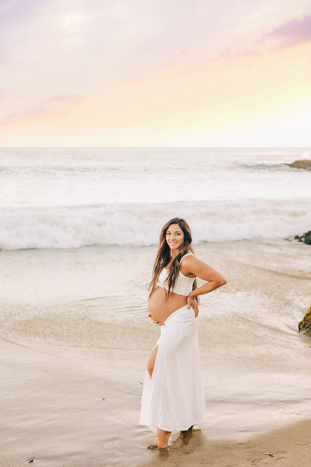 A mom to be walks on a beach at sunset in a white skirt and top with bump out after her 3D ultrasound in Long Beach