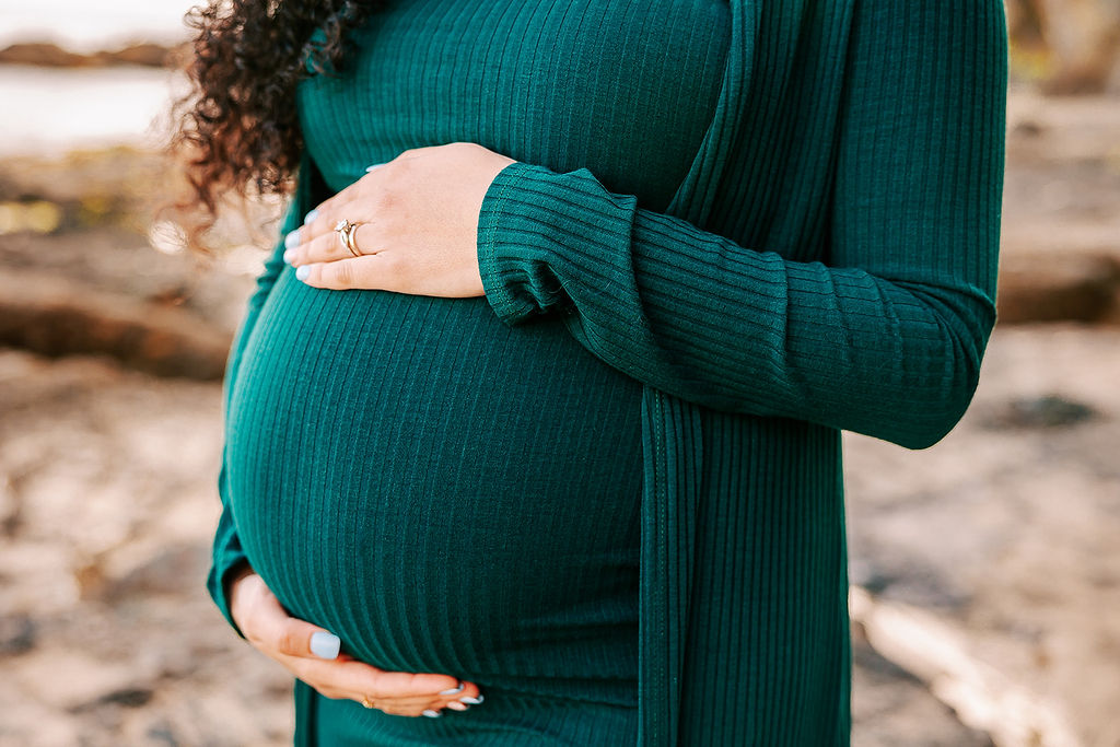 Details of a woman holding her pregnancy bump while standing on a beach in a green dress and sweater