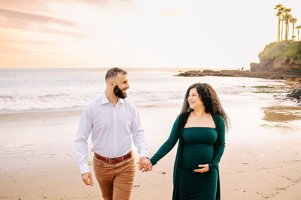 A smiling expecting couple walks while holding hands on a beach during sunset