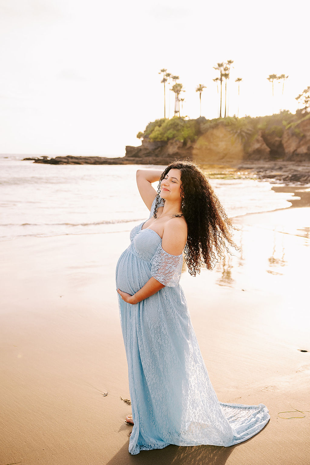 A smiling pregnant woman in a long blue lace maternity gown holds her hair back while walking on a beach at sunset after meeting a Doula in Long Beach