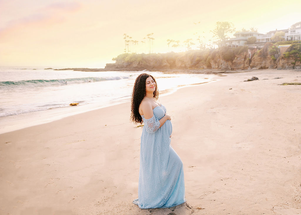 A smiling mom to be in a blue maternity dress walking on a beach at sunset after meeting a Doula in Long Beach