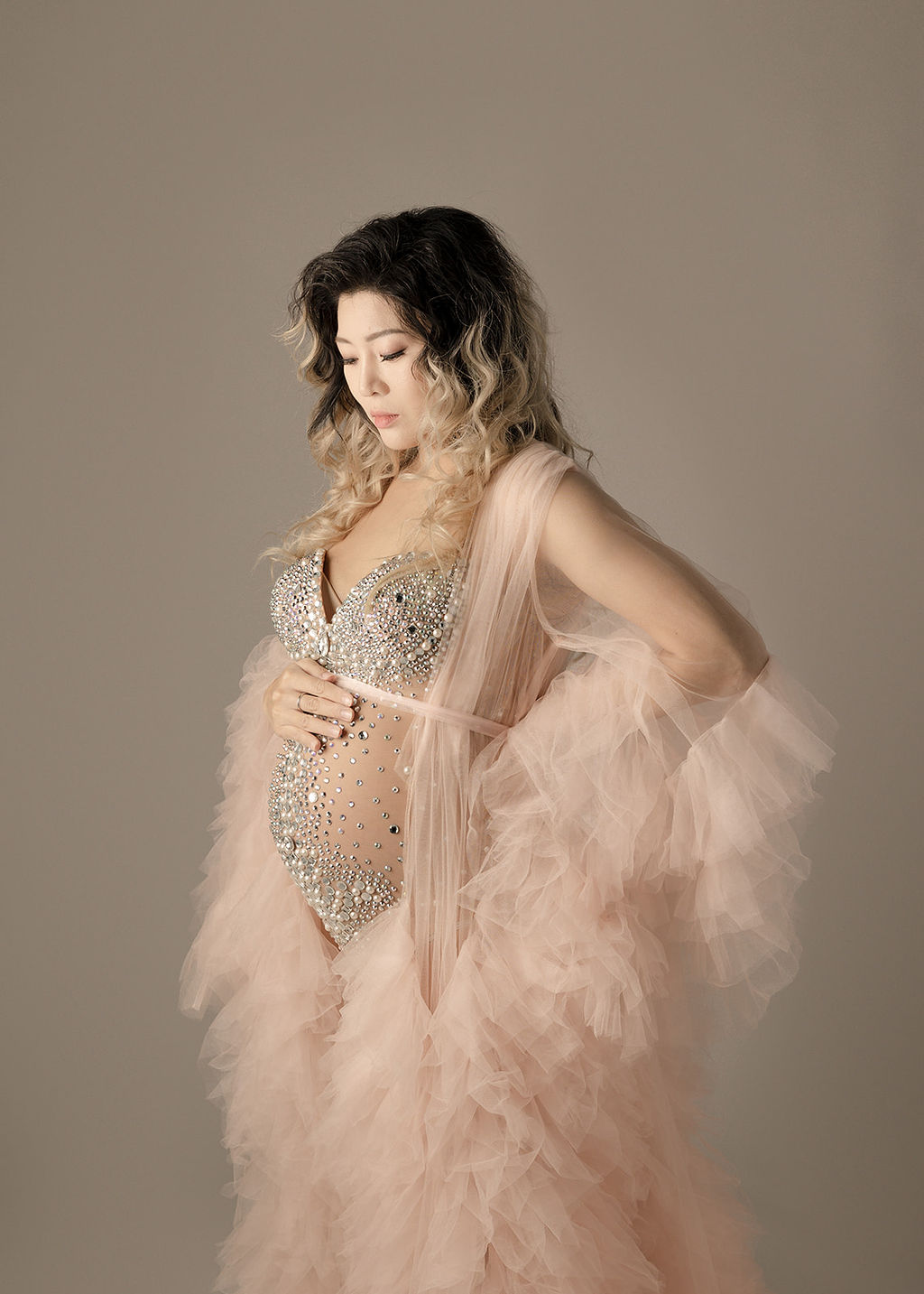 A mom to be stands in a pearl studded pink maternity gown looking down at her bump after meeting a Long Beach OBGYN